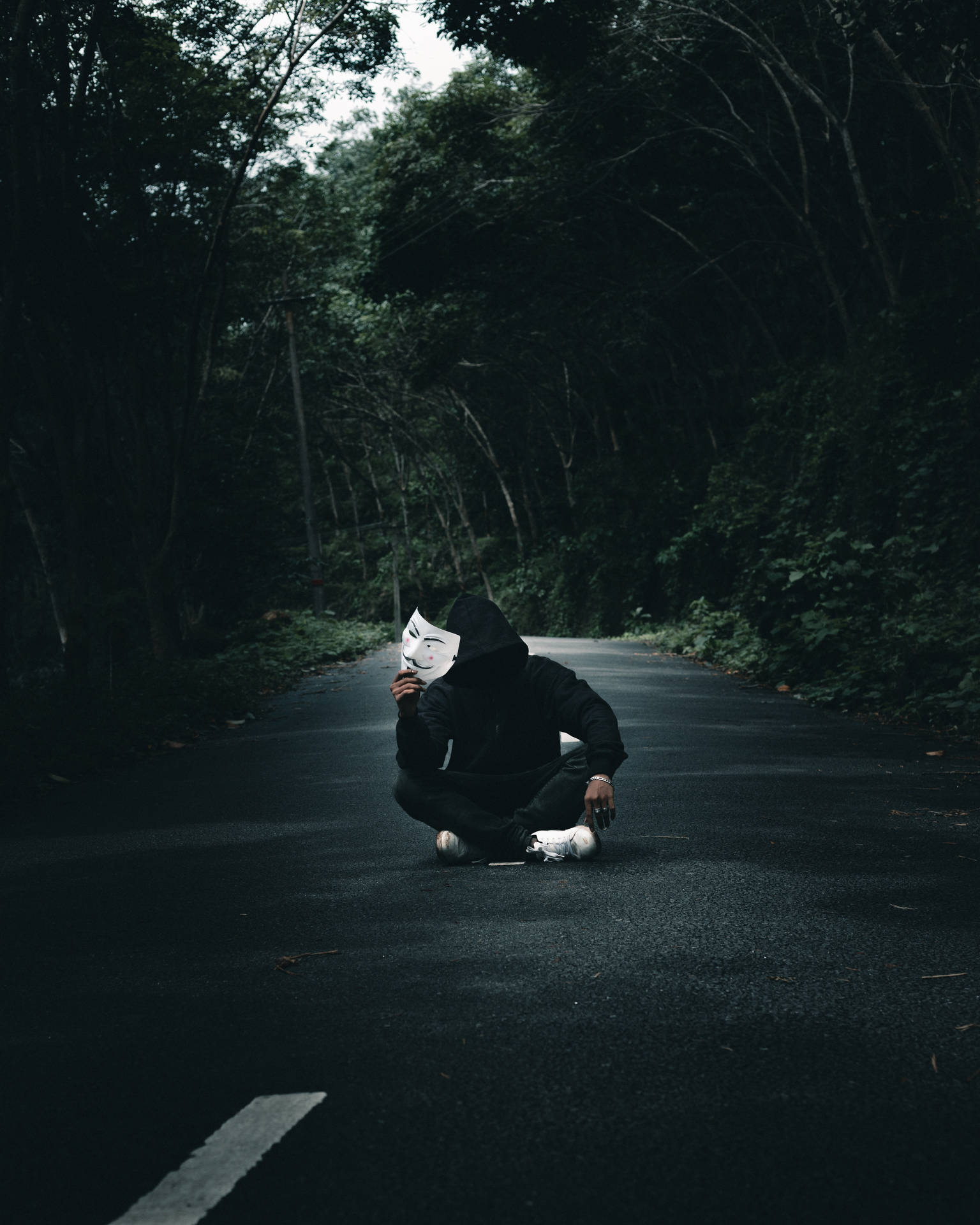 穿黑色連帽衫的人手持Anonymous面具在森林路上