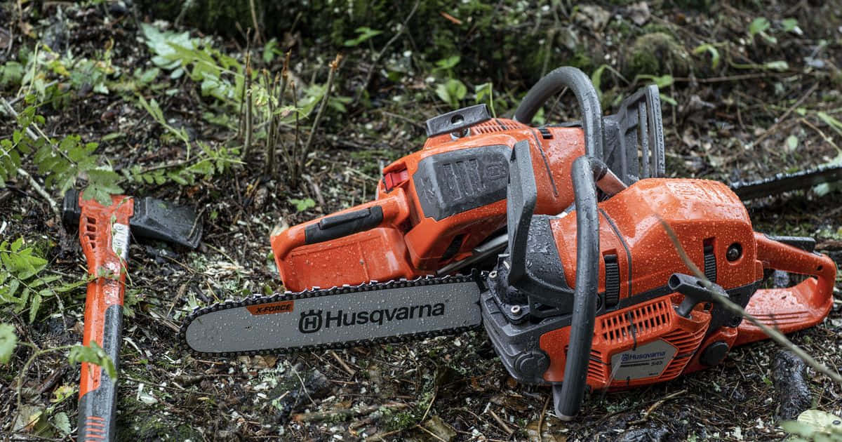 森林電鋸工具圖 壁紙 — 兩把橙色Husqvarna電鋸和一把斧頭濕漉漉地躺在森林地面上，周圍是綠色樹葉和落葉。
