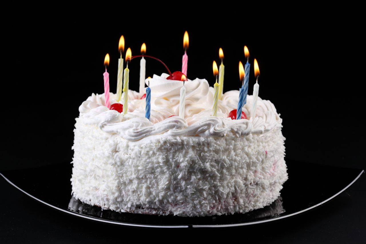 椰蓉裝飾生日蛋糕 壁紙 — 一個圓形白色椰子蛋糕，上面有燃燒的彩色蠟燭和櫻桃，放在黑色碟子上，背景深色。