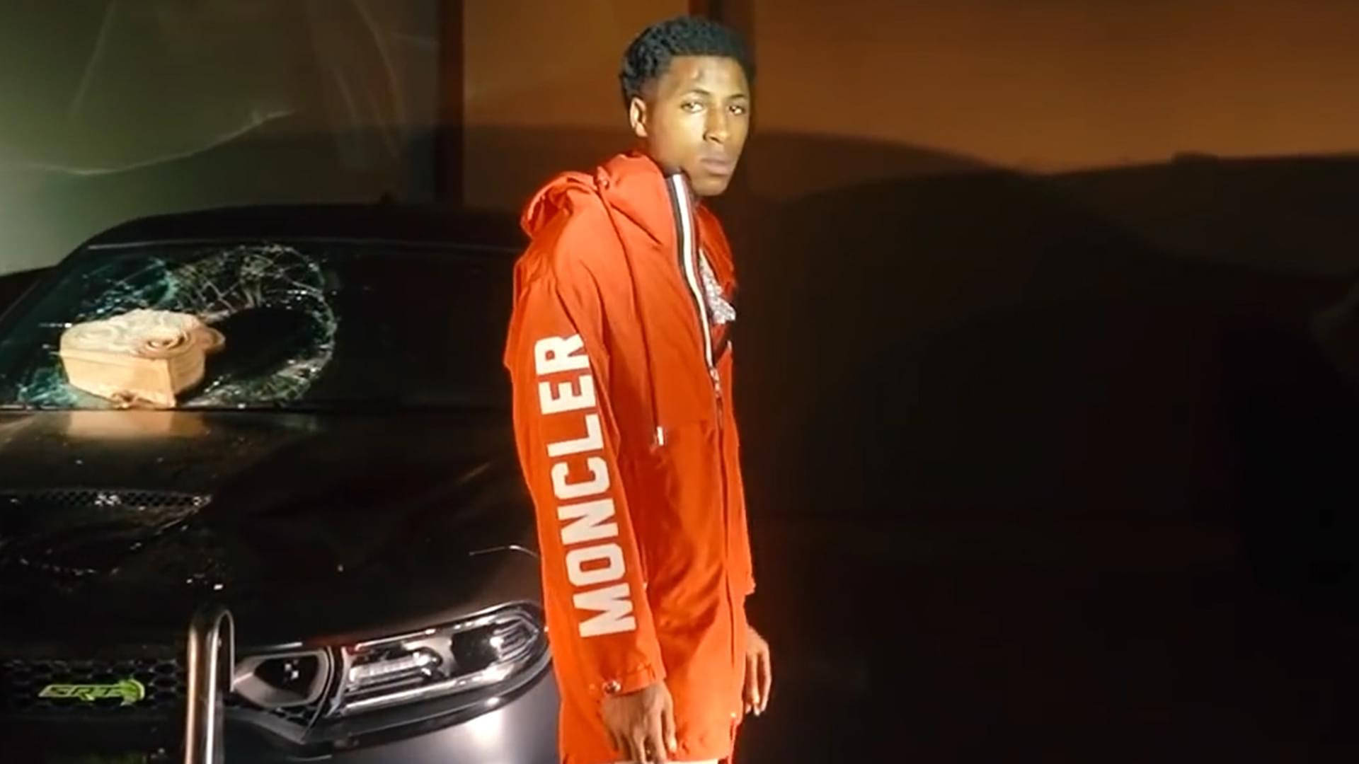 身穿橙色 Moncler 連帽衛衣的年輕男子在夜晚站在一輛被砸碎的黑色汽車旁。