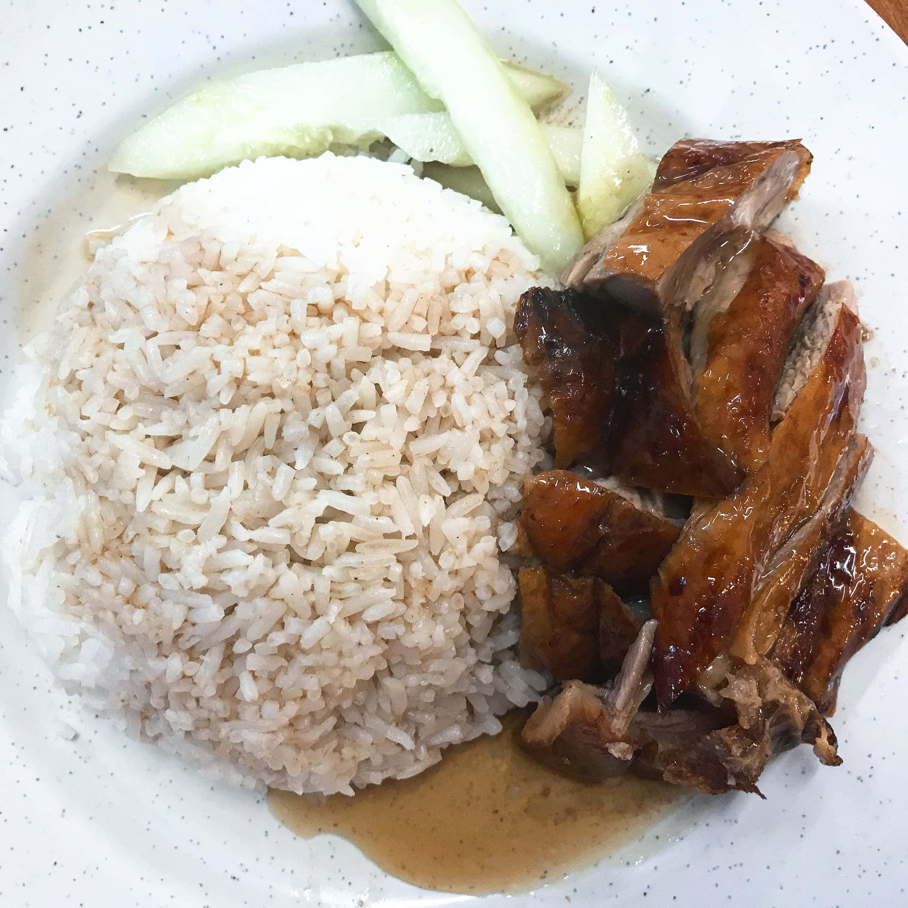 楊盛餐廳的鴨飯 壁紙 — 一碟帶有光澤的烤鴨，搭配糙米和青瓜片，放在有斑點的白色碟子上。