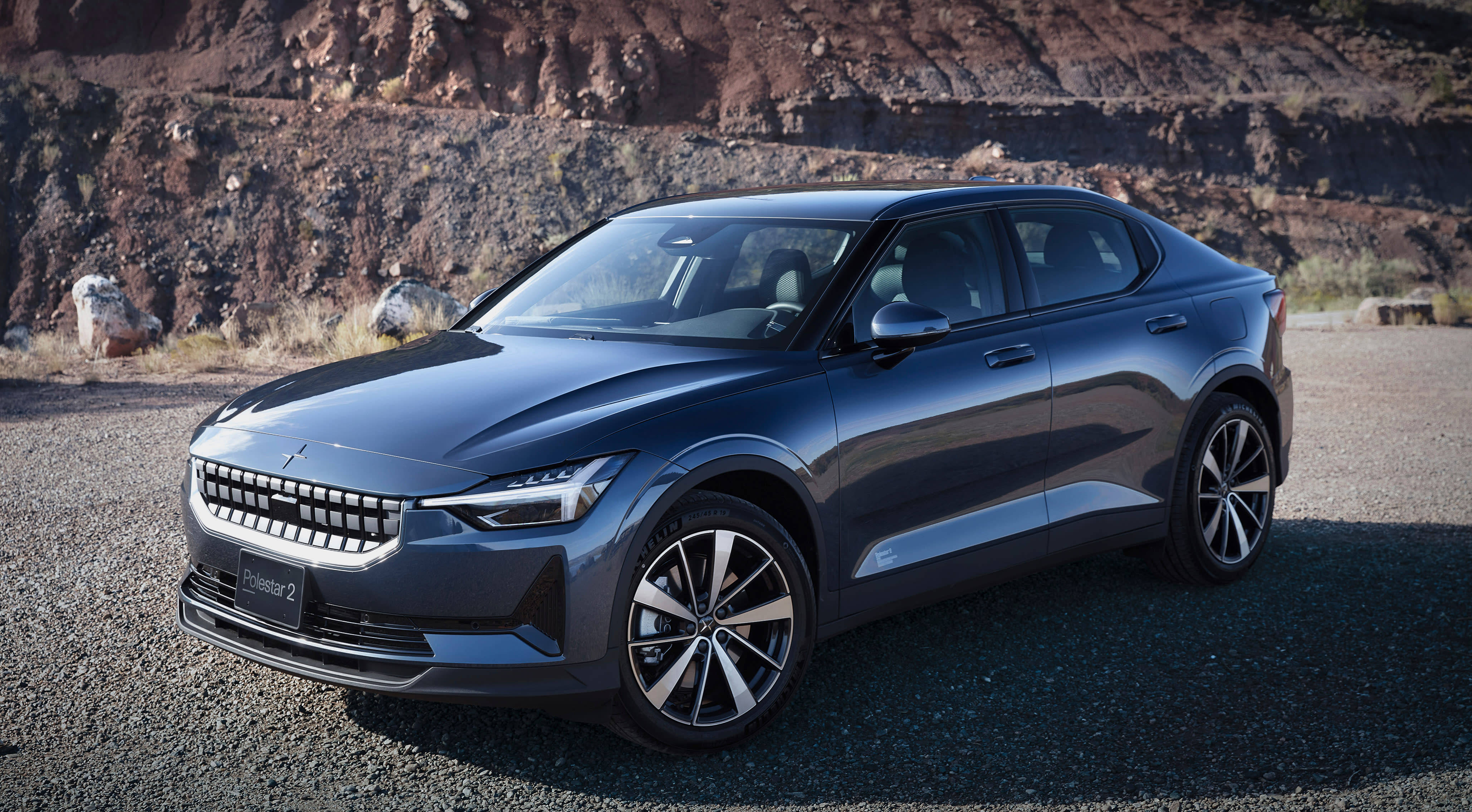 極星2於沙漠地點 壁紙 — 一輛時尚深灰色Polestar 2電動車停在碎石上，背景是崎嶇的紅色岩壁，在明亮的日光下。