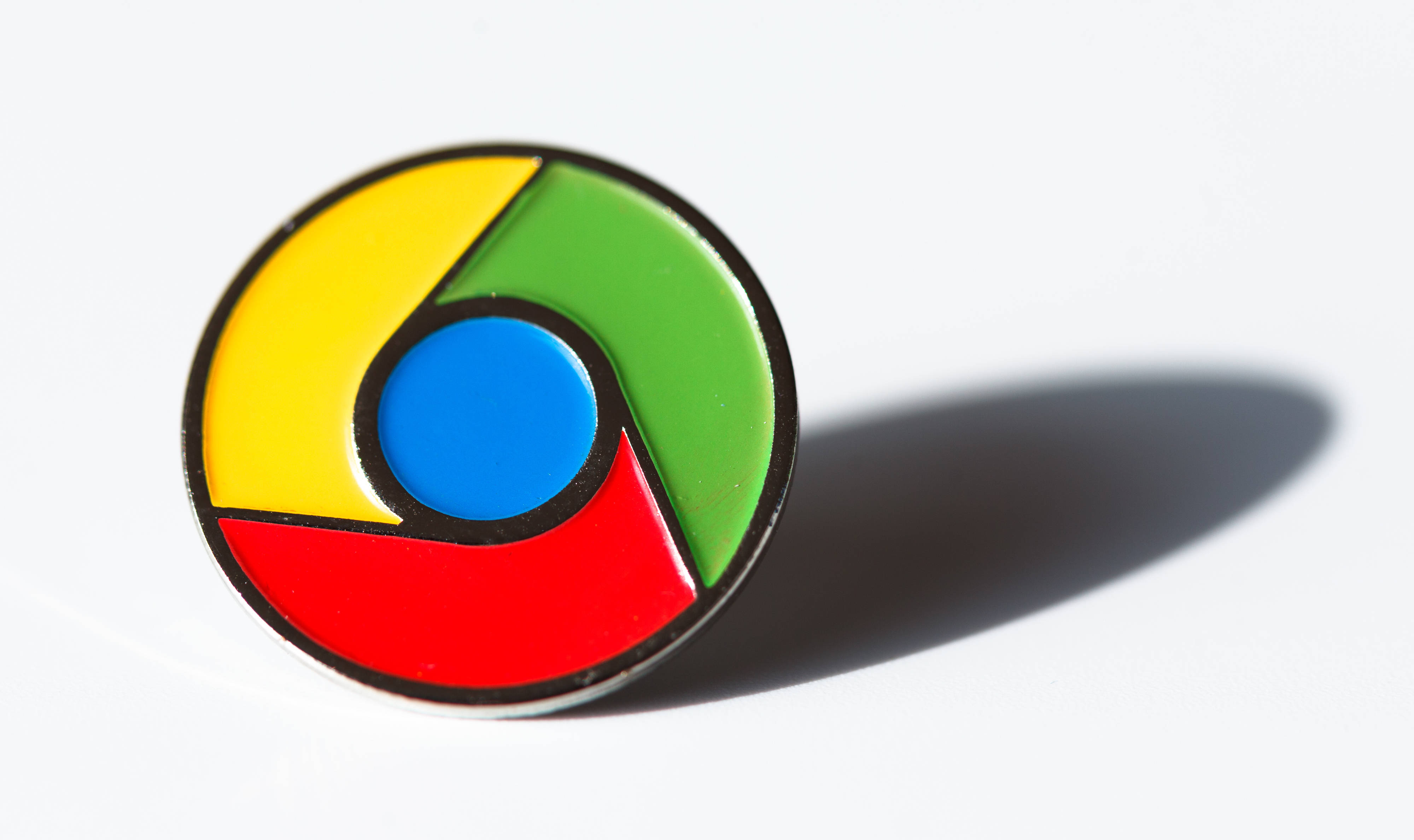 極簡Google Chrome胸針 壁紙 — 一個帶有黃色、綠色、紅色和藍色部分的光澤圓形 Chrome 標誌別針，在白色表面上投射出柔和陰影。
