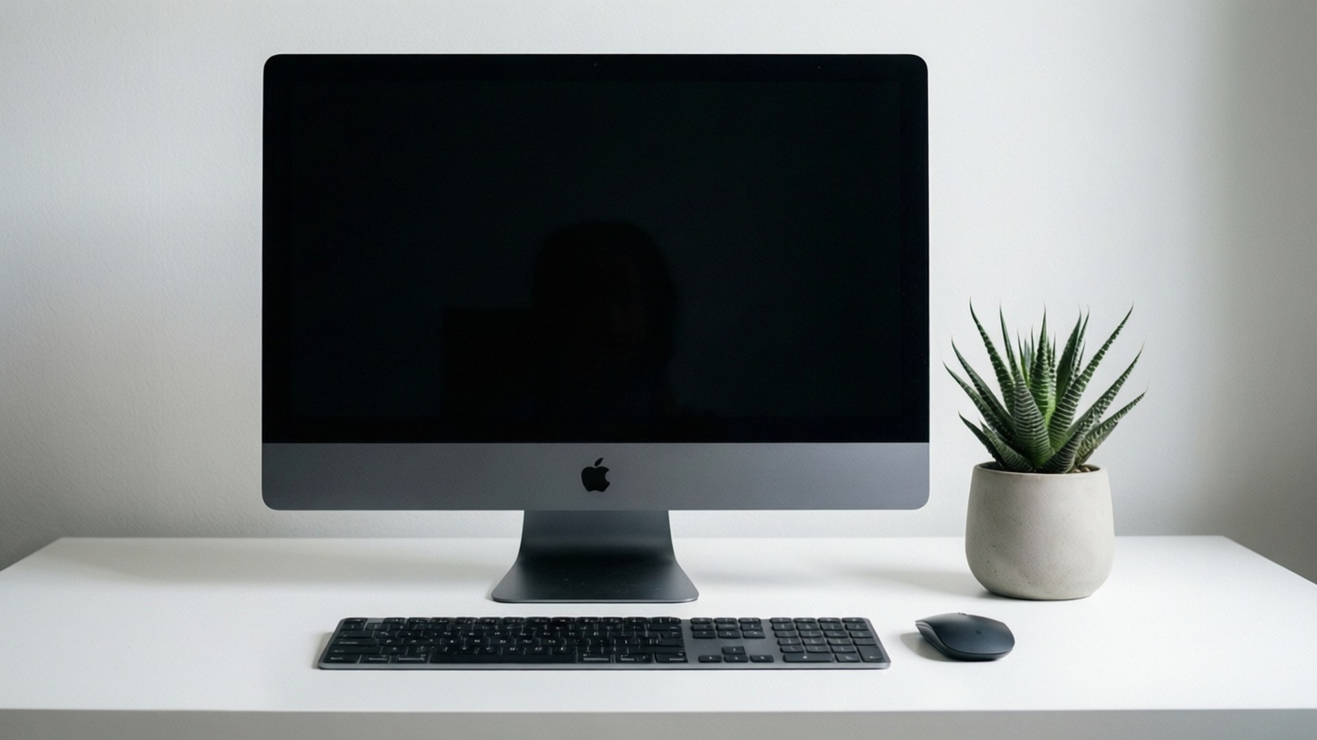 極簡iMac工作空間 壁紙 — 一部時尚的黑色 Apple iMac 放在白色桌子上，旁邊有一盆多肉植物和配套的鍵盤滑鼠。