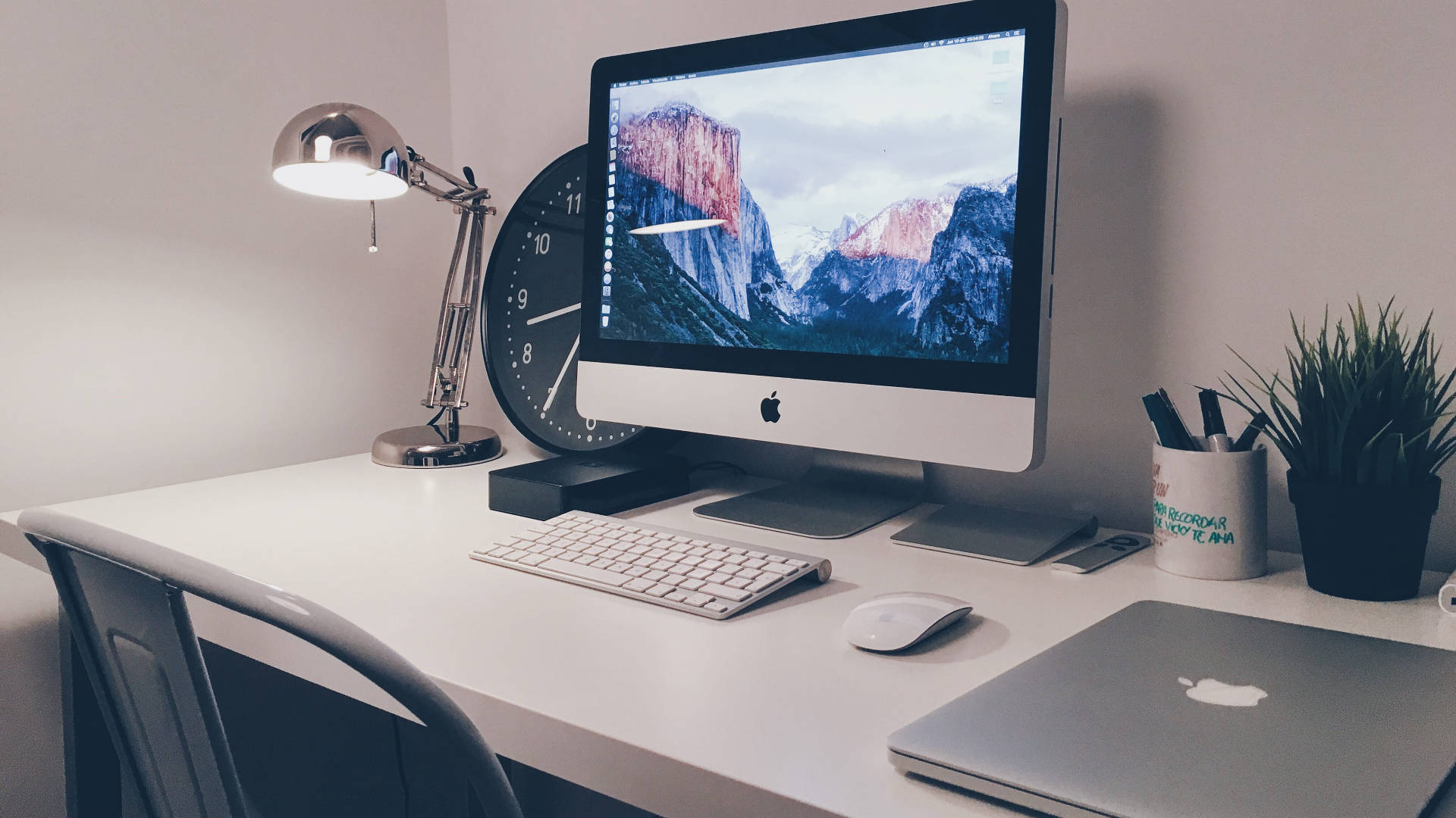 蘋果 桌布 壁紙 — 極簡白色桌面設置，配備 iMac 顯示山脈壁紙和銀色桌燈柔和發光。