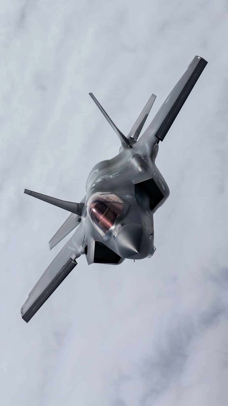 極簡風格F-35A飛行Android巨無霸飛機背景 壁紙 — 一架流線型灰色隱形戰鬥機在多雲的天空中翱翔，駕駛艙發出溫暖的光芒。