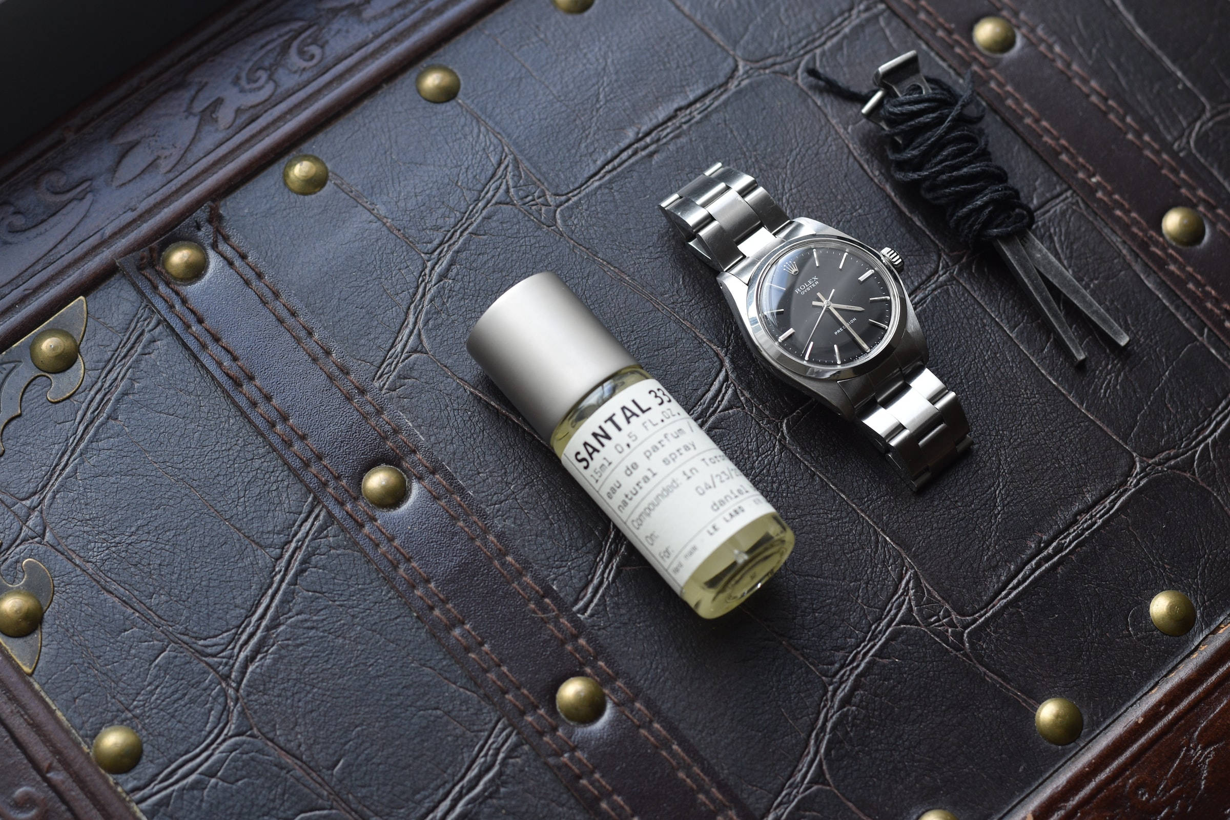 極簡風格勞力士 壁紙 — 一隻時尚的銀色手錶和一瓶 Santal 33 香水放在帶有黃銅鉚釘的紋理深棕色皮革表面上。
