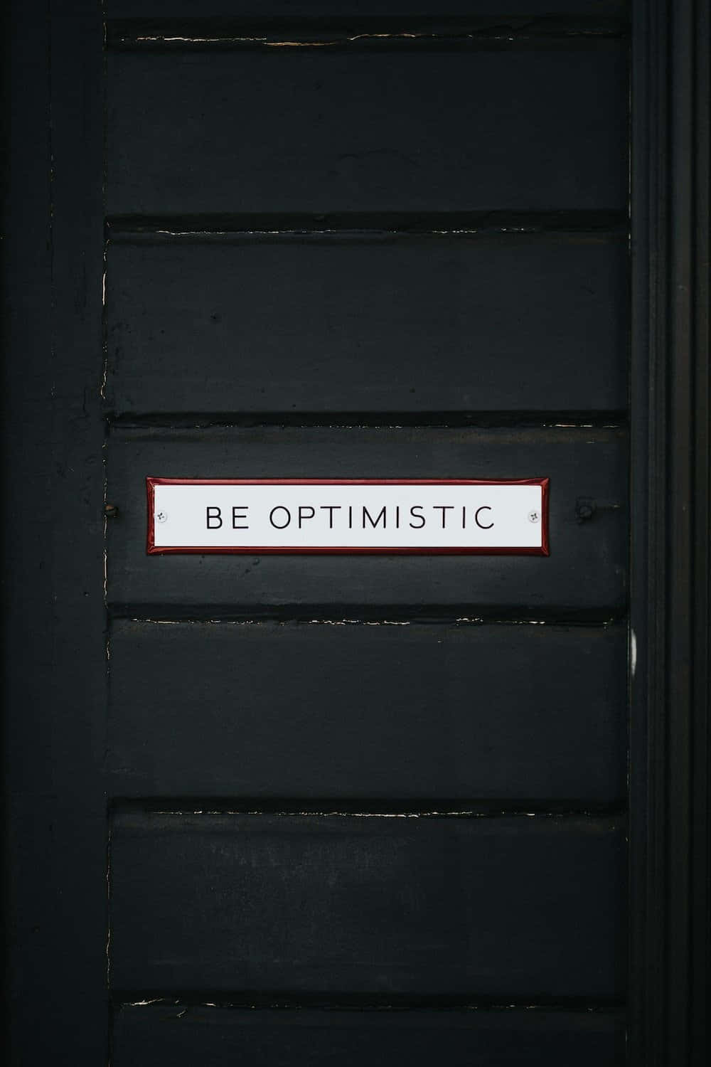 一扇黑色門，上面有一個白色標牌，寫著 be optimistic。