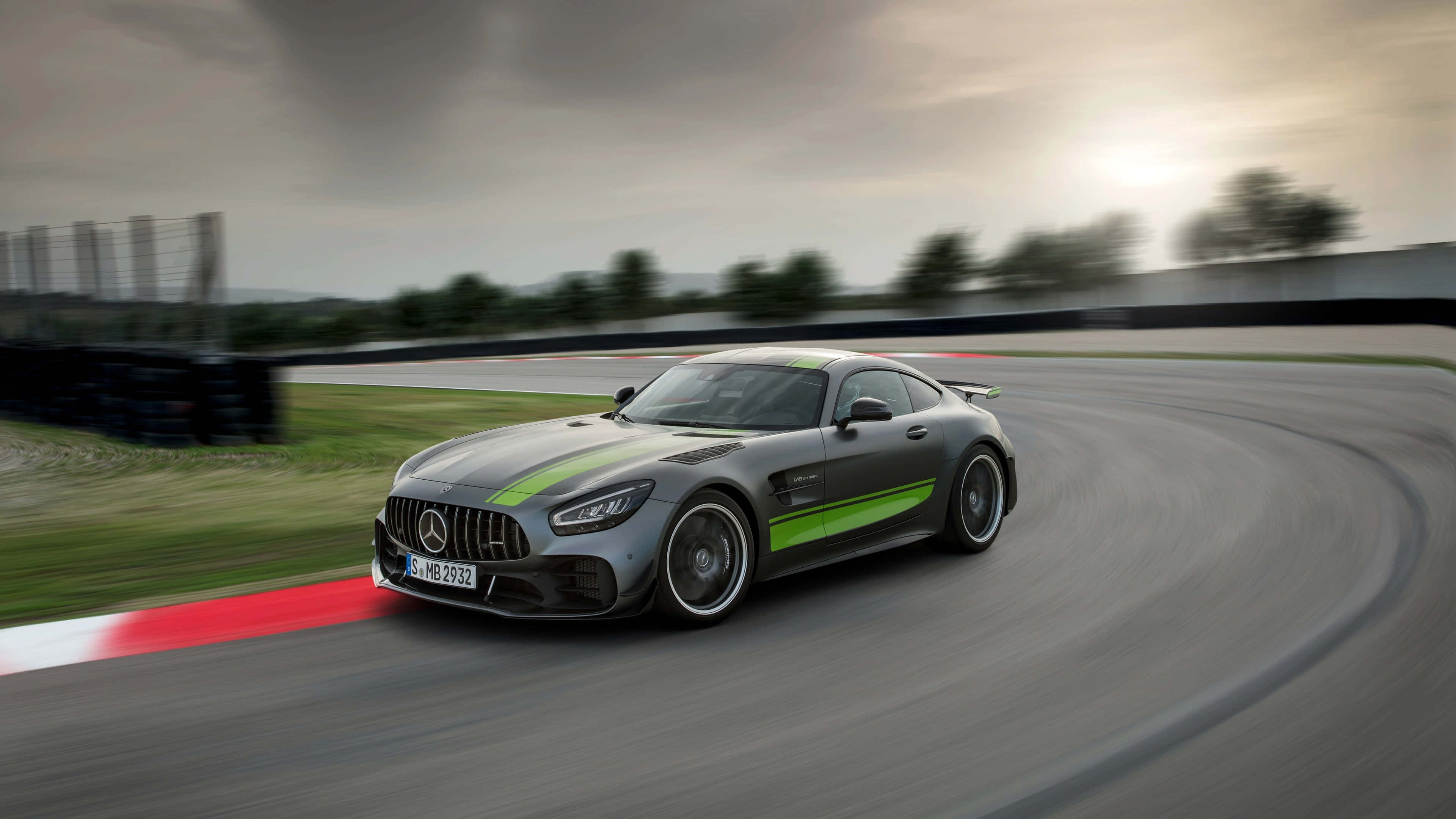 極致刺激 - AMG GT R 壁紙 — 一輛時尚的灰色Mercedes-AMG GT賽車，帶有綠色條紋，在多雲的天空下沿著彎曲的賽道飛馳。