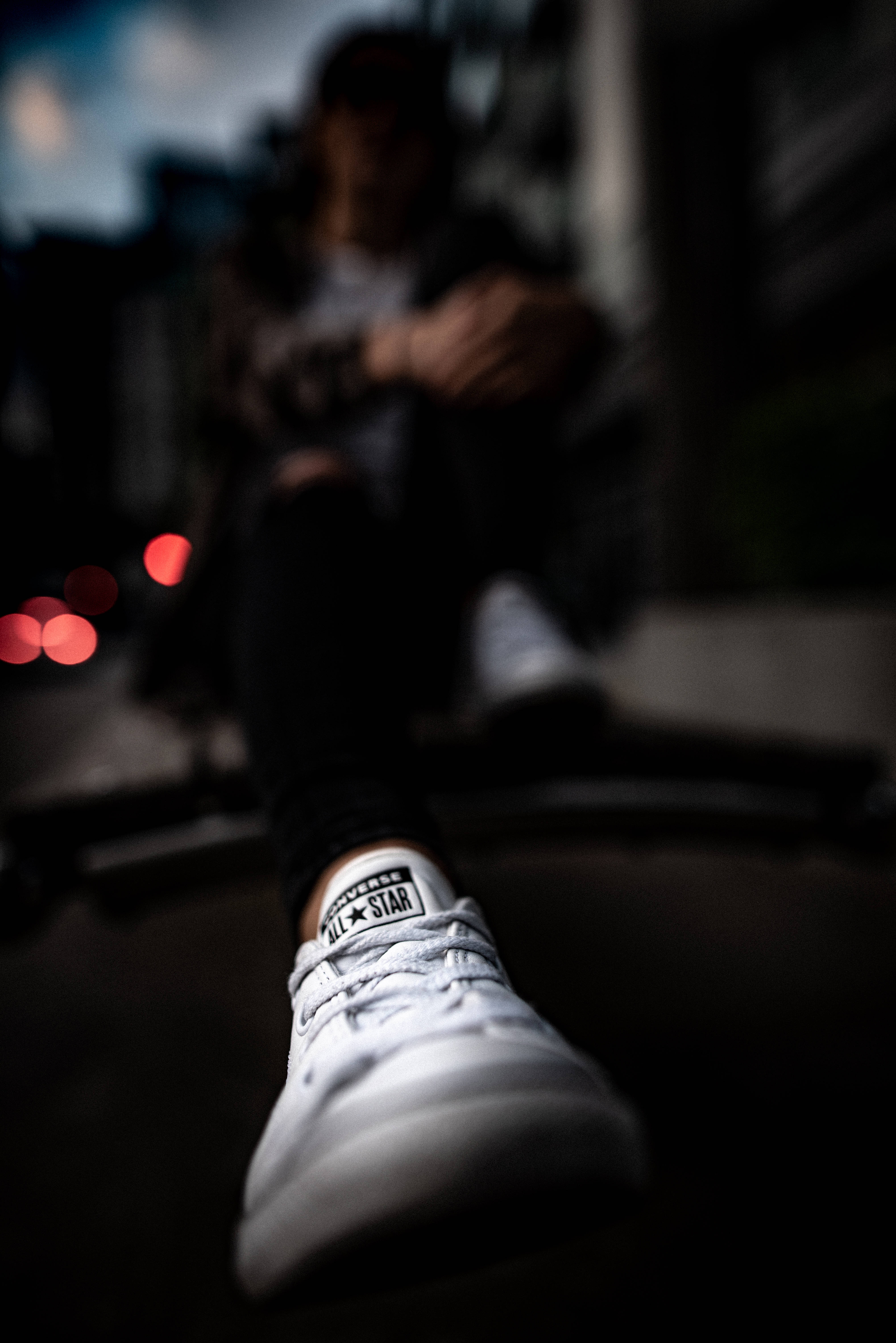 極致聚焦的白色Converse鞋 壁紙 — 低角度拍攝聚焦於一雙白色 Converse 運動鞋，背景是模糊的人坐在昏暗的城市環境中。