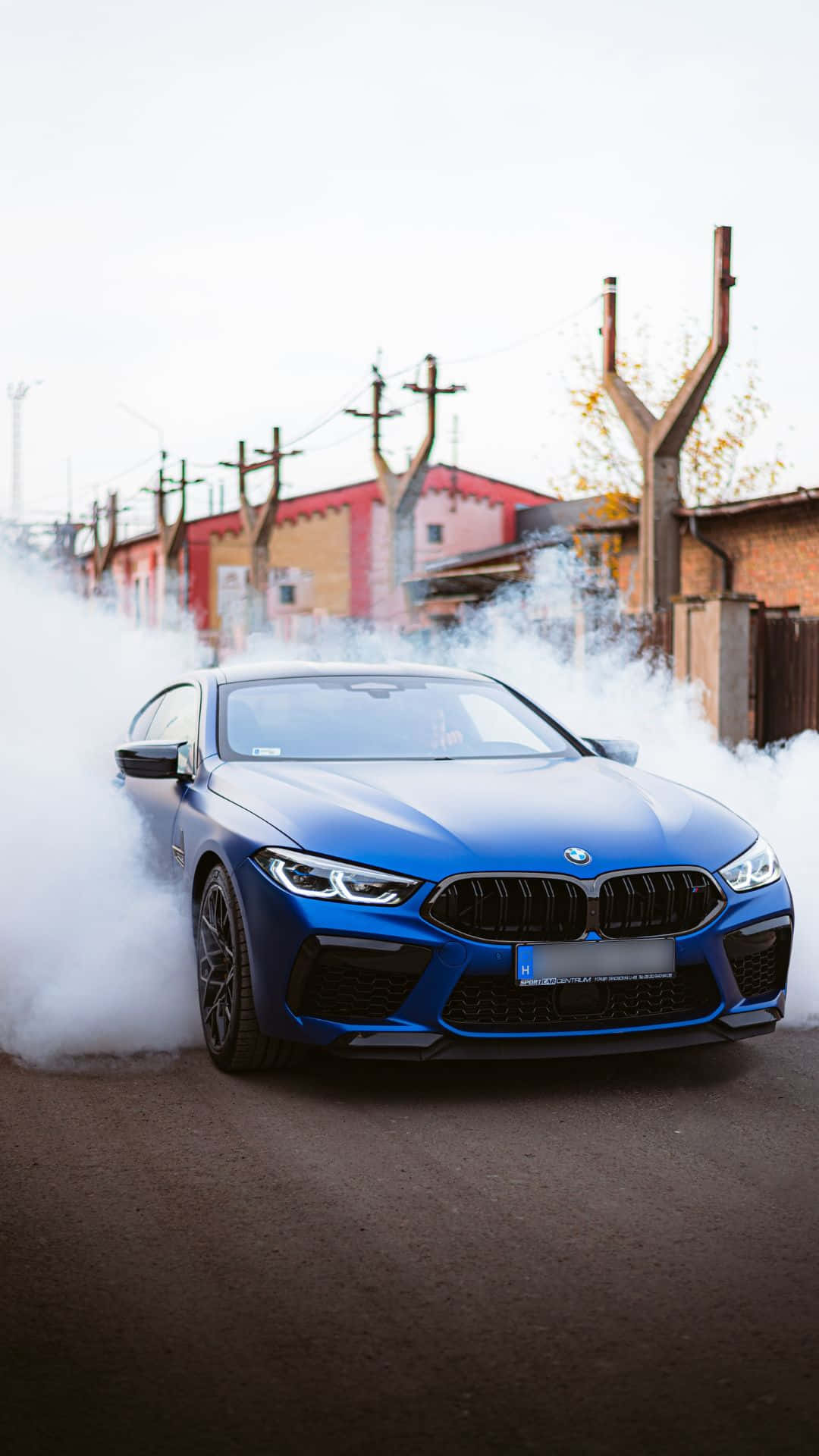極致駕駛體驗：BMW內建安卓系統 壁紙 — 一輛充滿活力的藍色BMW M8在濃煙中飄移，背景是工業建築的都市街道。