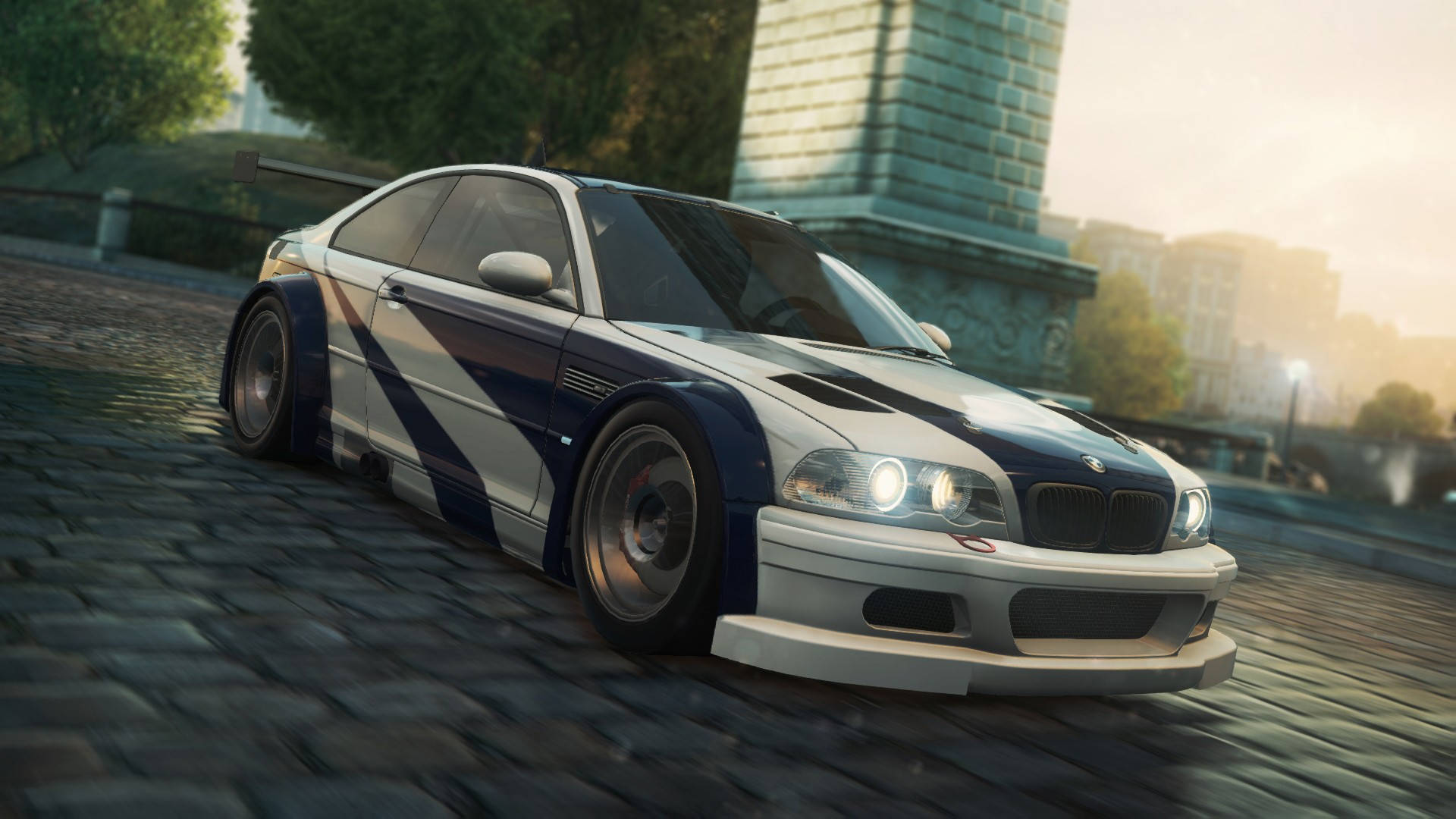 《極速快感》BMW M3 GTR E46 壁紙 — 一架白色和藍色的BMW M3賽車在鵝卵石街道上飛馳，背景有一座石頭紀念碑。