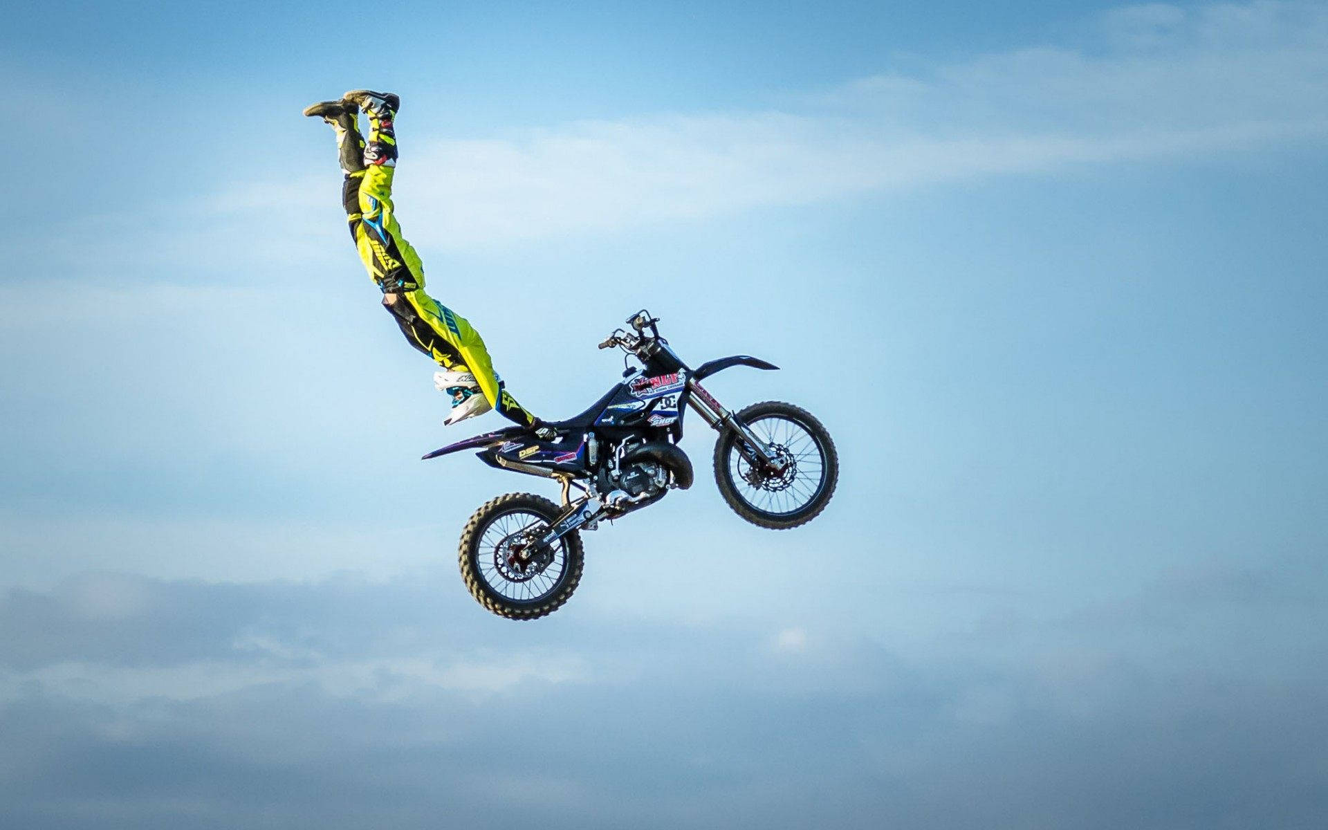 極限運動 FMX 自行車跳躍 壁紙 — 一名身穿亮黃色裝備的越野電單車手，在佈滿雲朵的藍天下，與他的電單車一起表演倒立空中特技。