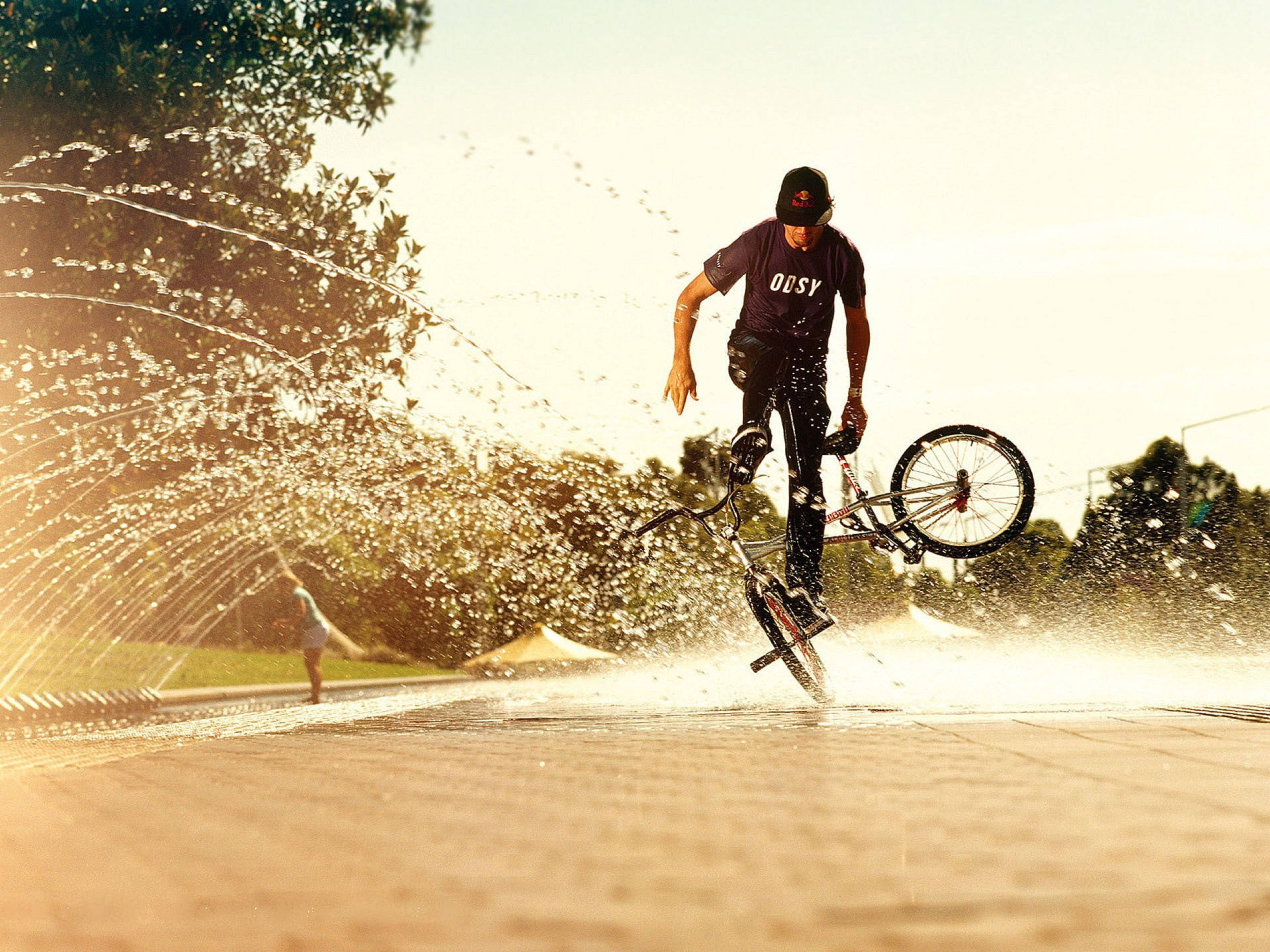 極限運動BMX水上特技 壁紙 — 一名BMX車手在陽光明媚的公園中，於噴灑金色水花的噴泉上方表演空中特技。
