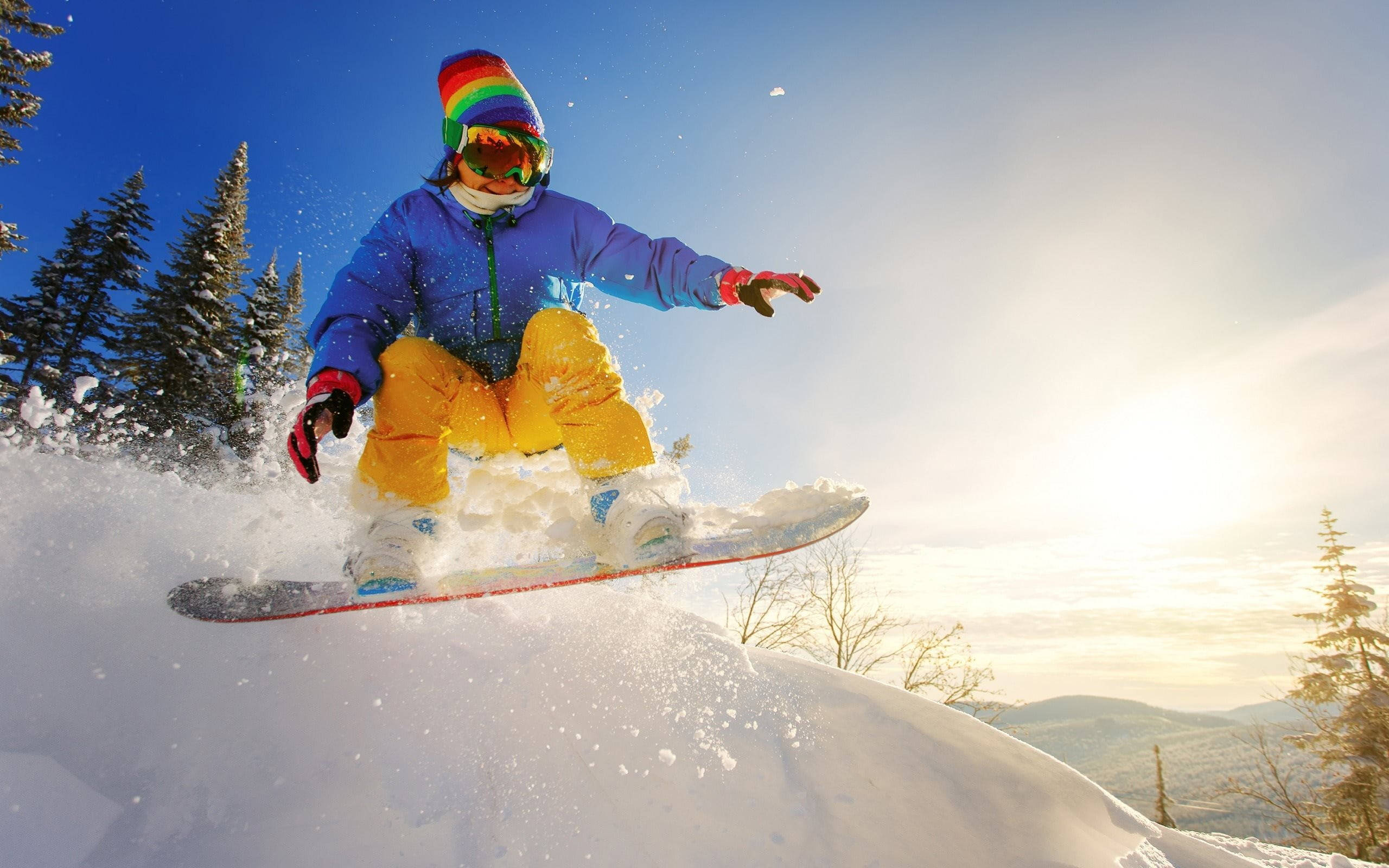 極限運動滑雪山頂 壁紙 — 一個穿著色彩繽紛裝備的小孩在藍天白雲下，於松樹和山巒環繞的雪坡上空玩滑雪板。
