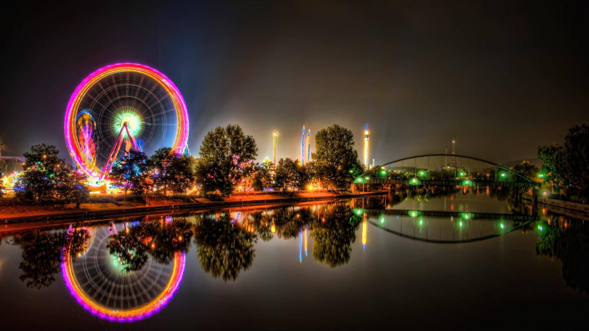 樂園夜景燈光 壁紙 — 遊樂園中平靜水面上倒映著色彩繽紛的摩天輪和橋樑的充滿活力的夜景。
