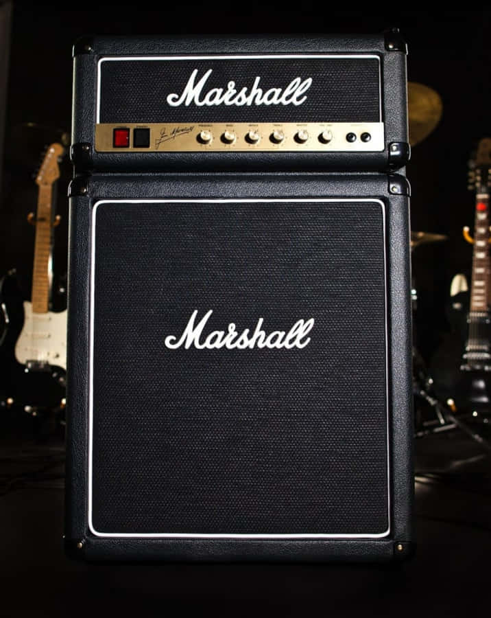 樂手使用經典管狀擴音器演奏電吉他 壁紙 — 一個黑色Marshall結他擴音器堆疊，帶有白色標誌和旋鈕，在昏暗燈光下與結他一起站在舞台中央。