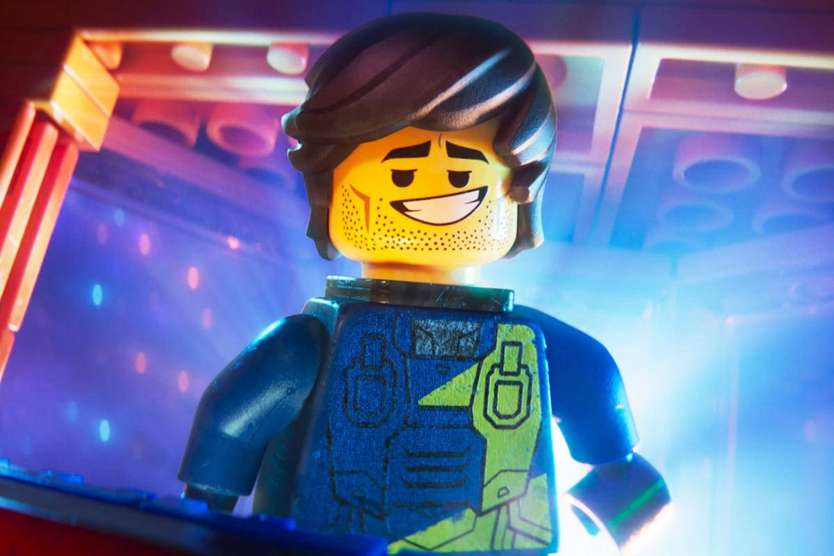 《樂高大電影2》雷克斯·丹格維斯特 壁紙 — 一個微笑的LEGO迷你人偶，棕色頭髮和藍色背心，站在充滿活力的霓虹燈舞台場景中。