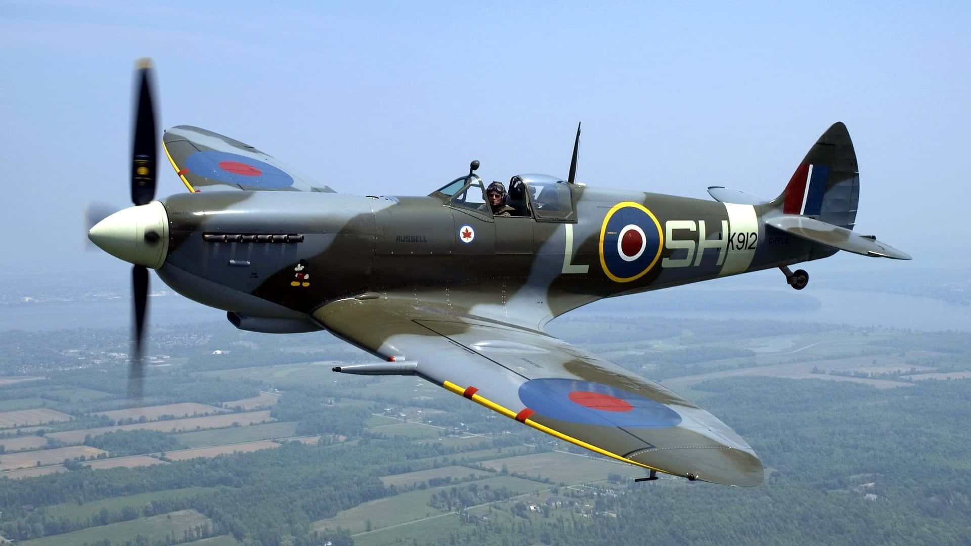 標誌性的英國戰鬥機——Supermarine Spitfire 壁紙 — 一架復古英國噴火戰鬥機在晴朗藍天下翱翔於拼布般的鄉村上空。