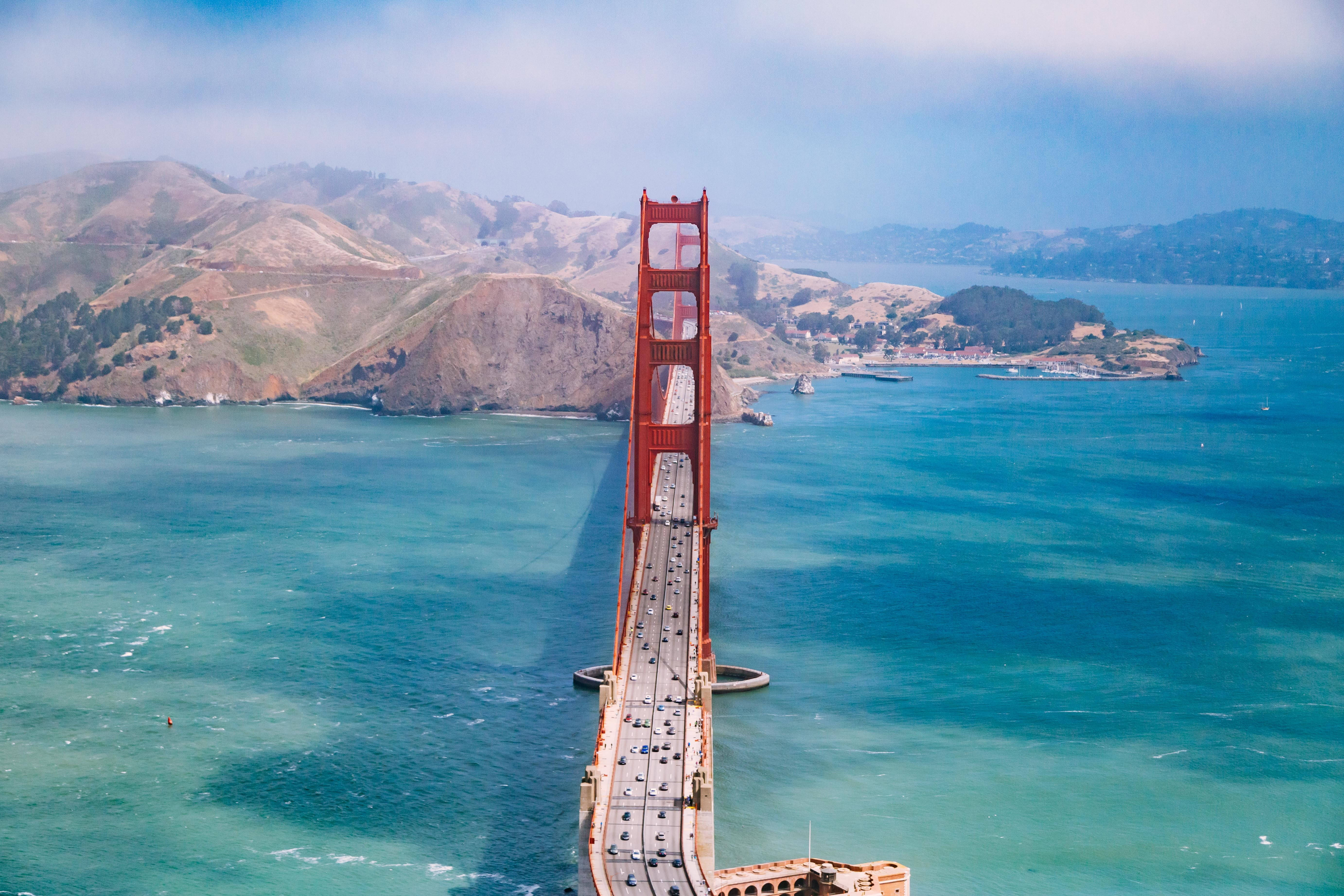 San Francisco Phone 桌布 壁紙 — 標誌性紅色金門大橋橫跨碧綠海水嘅航拍視角，背景有朦朧山丘。