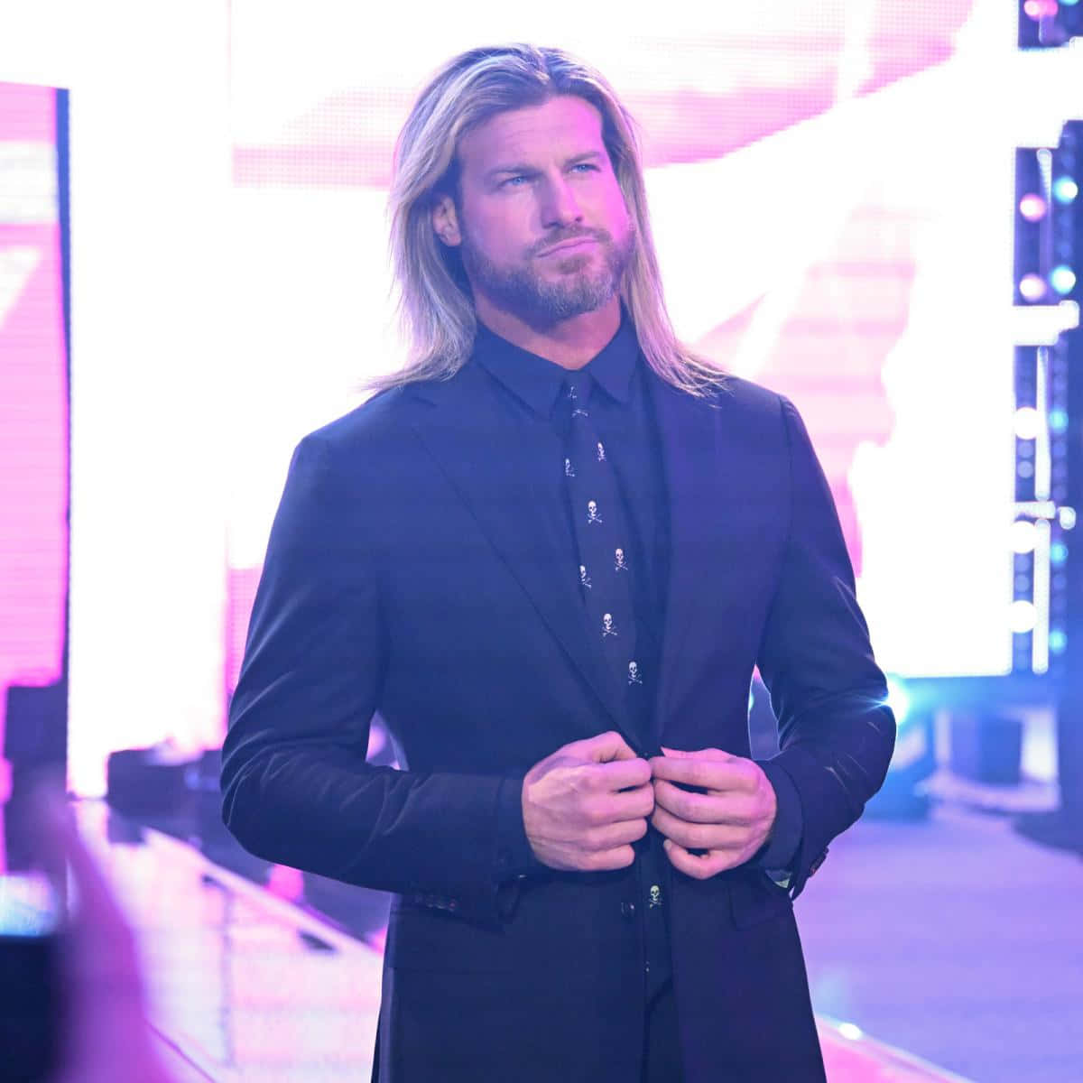 標題：Dolph Ziggler - WWE超級巨星動態演出 壁紙 — 一名留著金色長髮和鬍鬚的男子身穿黑色西裝，在充滿活力的粉紅色和紫色舞台燈光下扣上外套鈕扣。