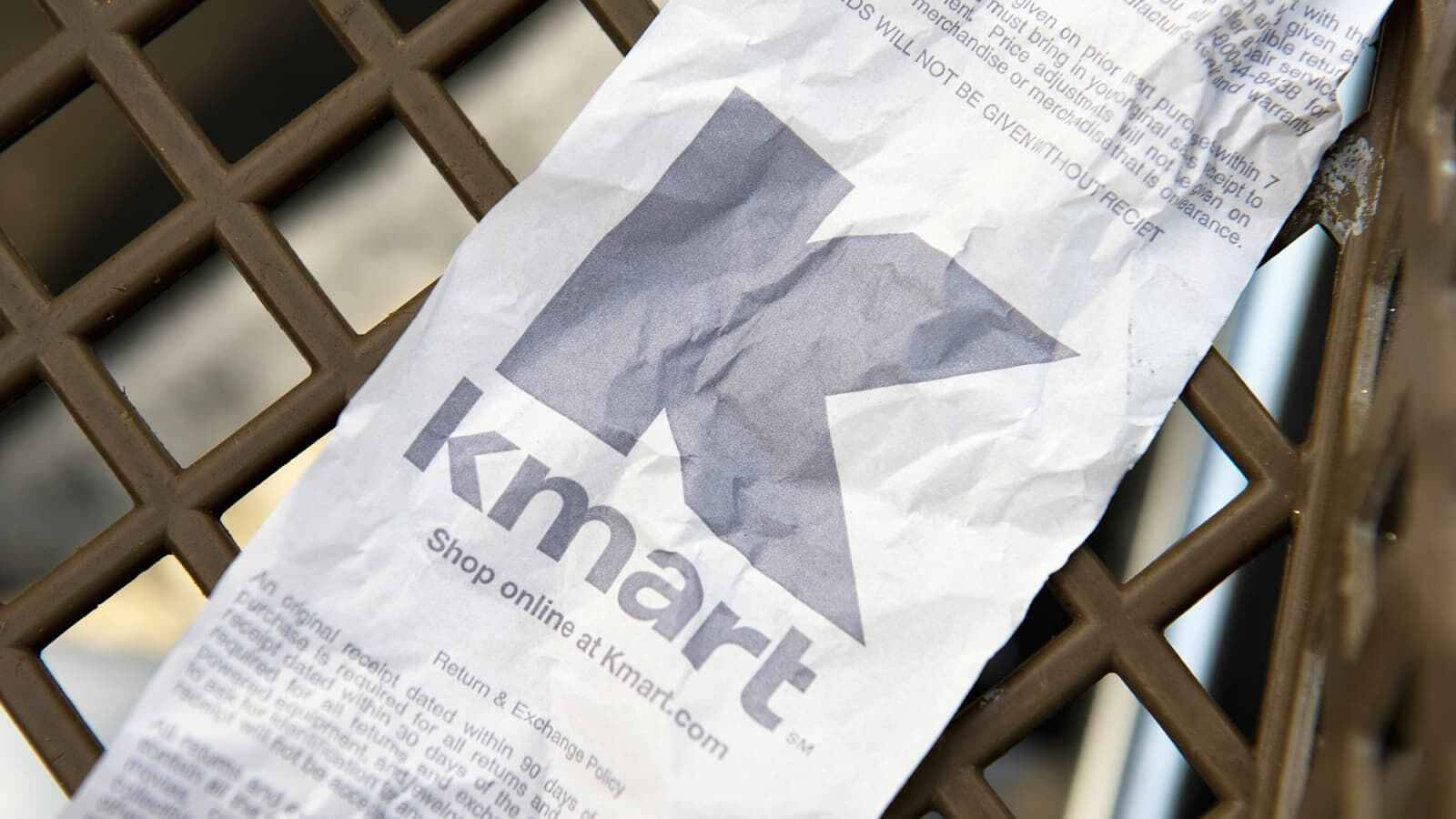 標題：Kmart購物收據 壁紙 — 一張皺巴巴的Kmart收據斜放在棕色塑膠購物車格柵上。