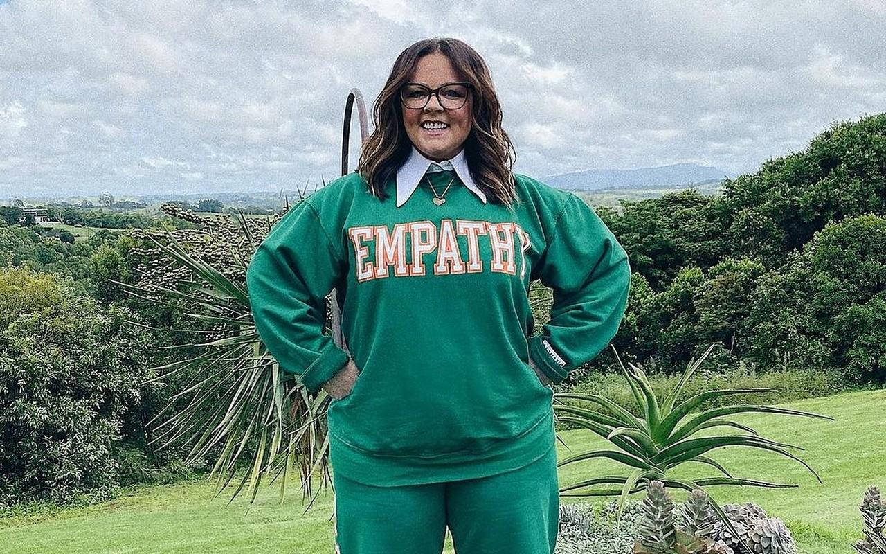 穿著綠色 'EMPATHY' 運動衫的女性在戶外長滿青草的山丘上微笑，背景有樹木和山脈。