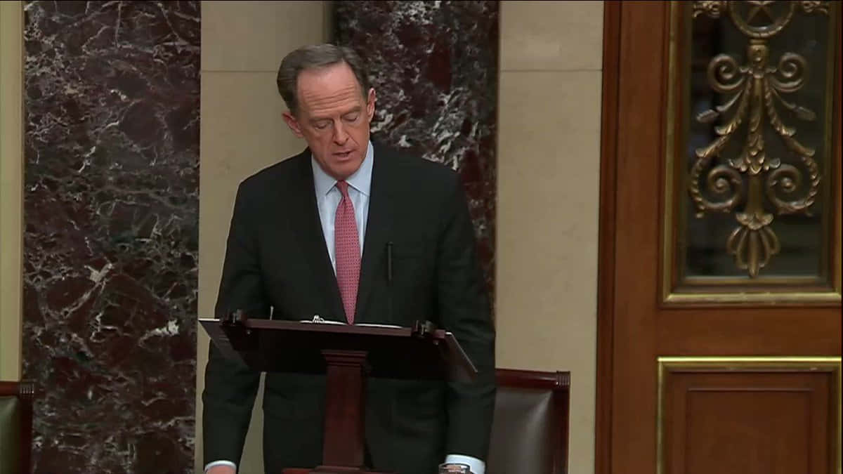 標題：參議員帕特·圖米在參議院大廳 壁紙 — 一位身穿深色西裝和紅色領帶的男子在宏偉的大理石和木鑲板房間的木製講台上發言。