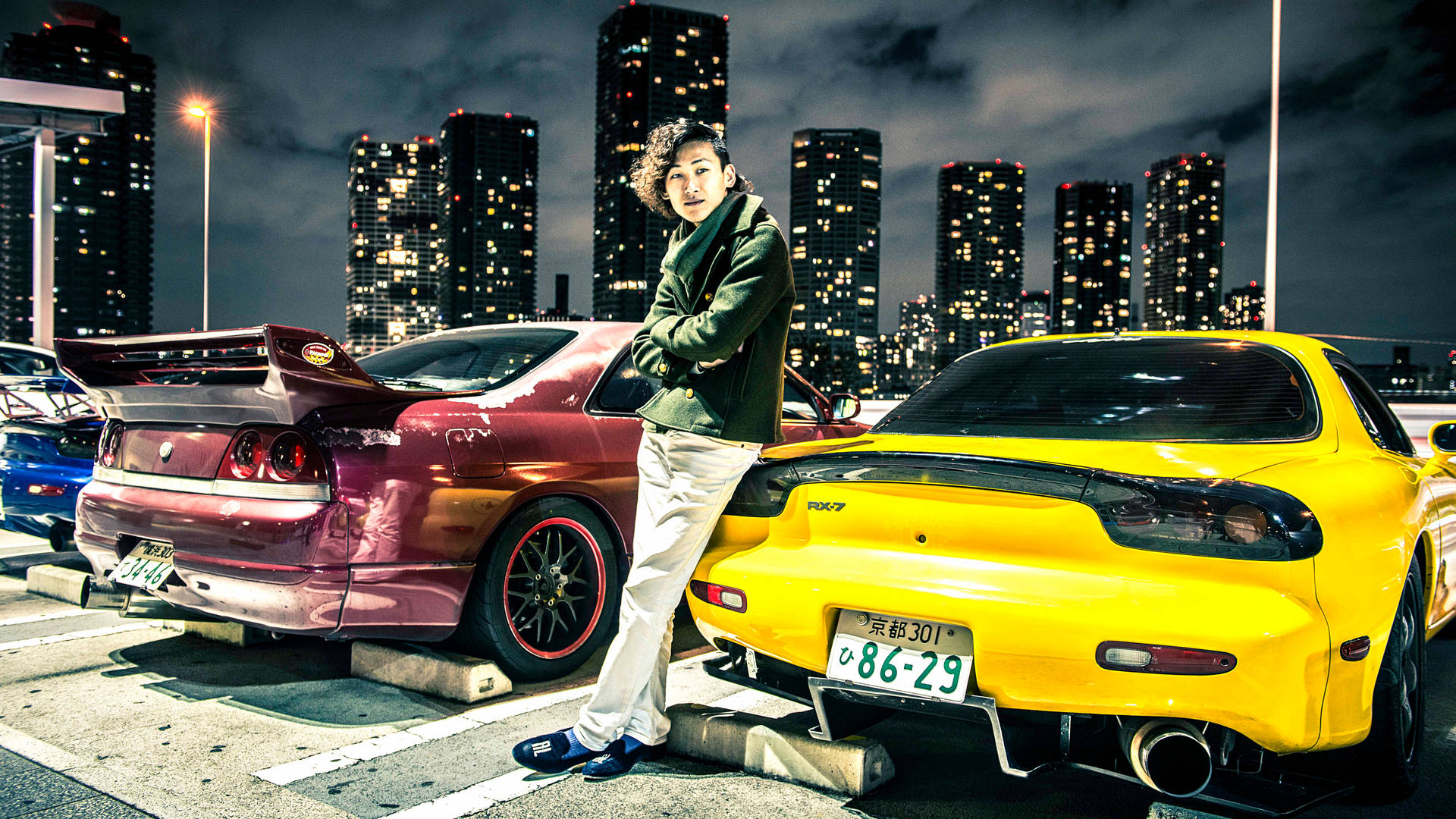 年輕男子在夜晚的城市停車場中靠在兩輛跑車之間。