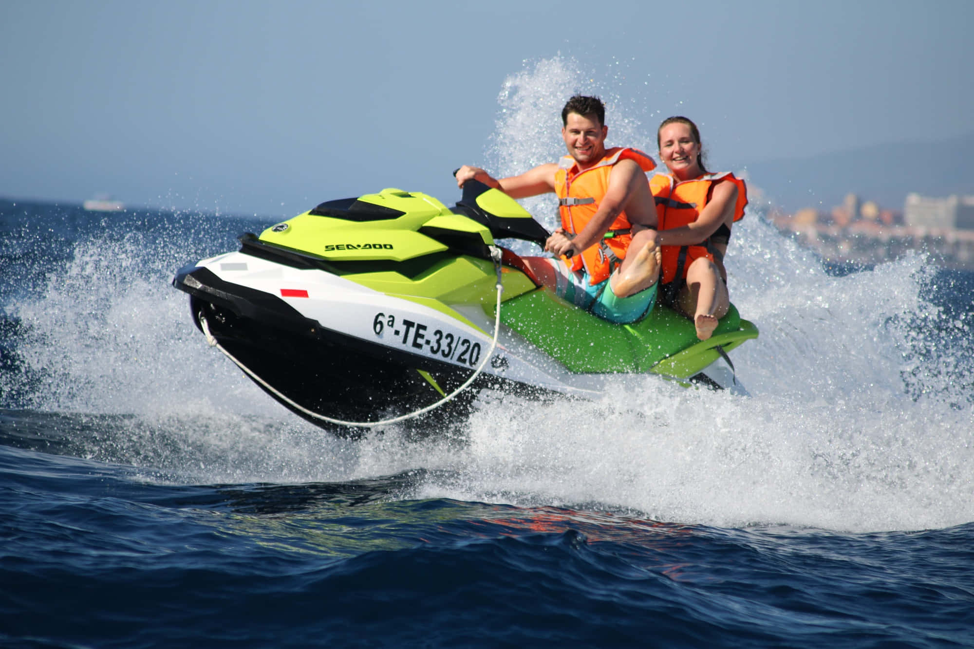 標題：熱愛刺激的夫婦在清澈水域加速駕駛摩托艇 壁紙 — 一對微笑的情侶騎著亮綠色的水上摩托車，在波濤洶湧的藍色海面上留下白色水花痕跡。