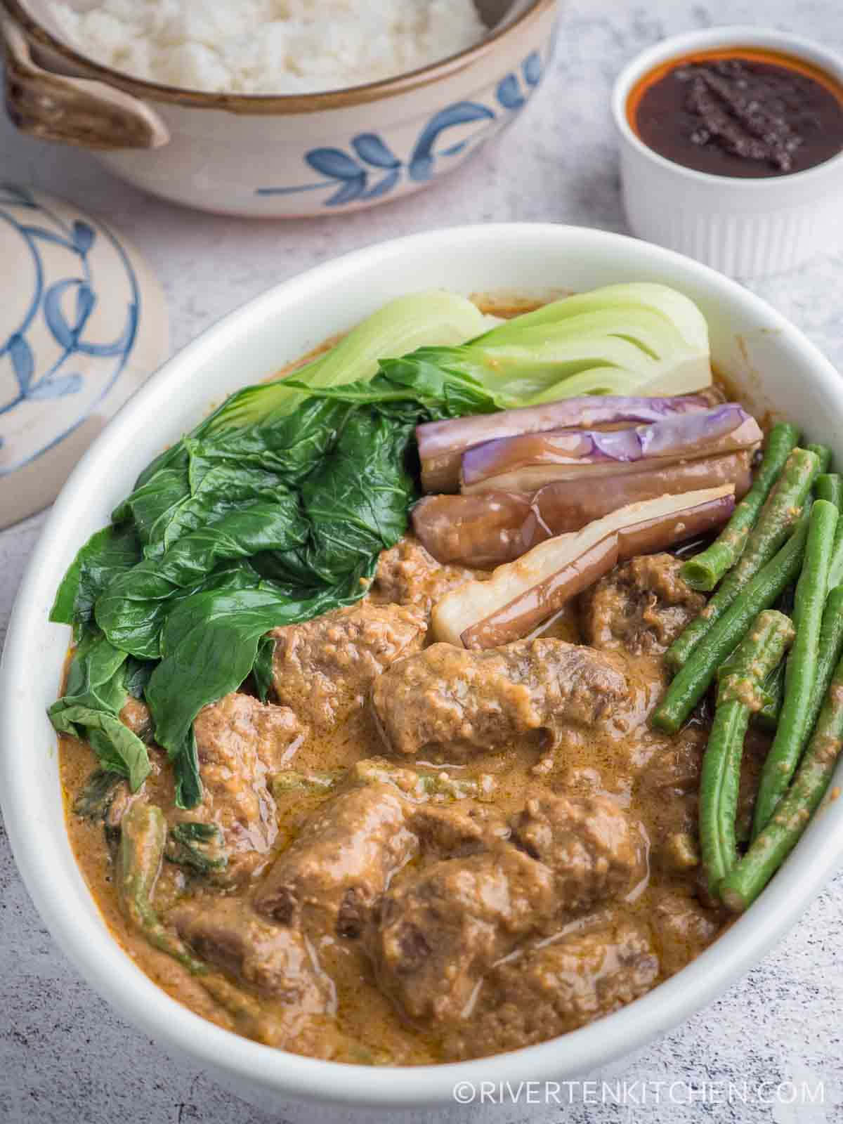 標題：美味的菲律賓菜式 Kare-Kare 壁紙 — 一碗熱氣騰騰的濃郁棕色咖喱，內有嫩肉、青豆、白菜和茄子，配上白飯和蘸醬。