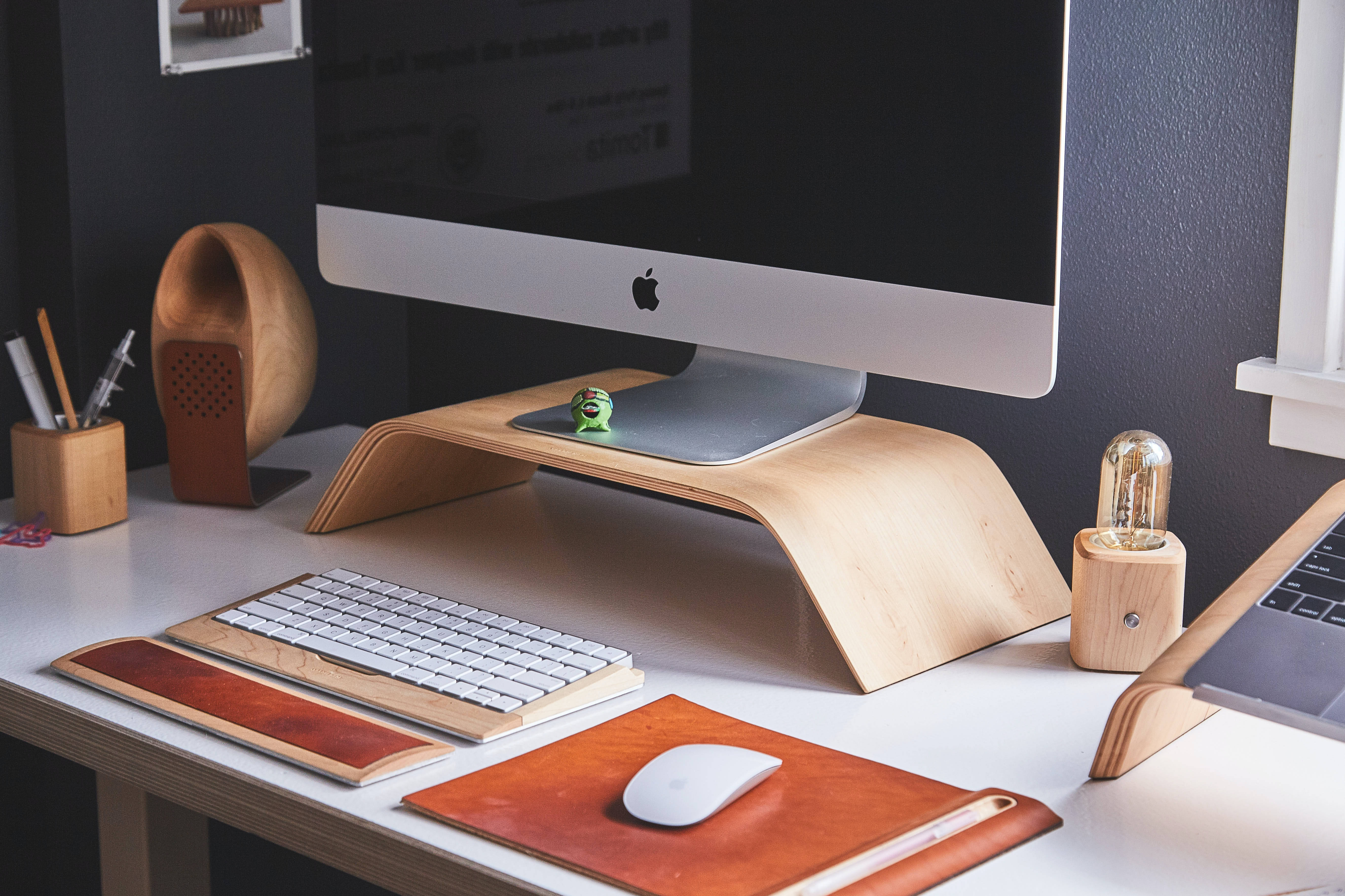 標題：美學和諧 - 搭配咖啡的 Mac 工作區 壁紙 — 一個簡約現代的辦公桌設置，配有木製支架，放置 iMac 鍵盤、滑鼠和溫暖的皮革配件，在柔和環境光下。