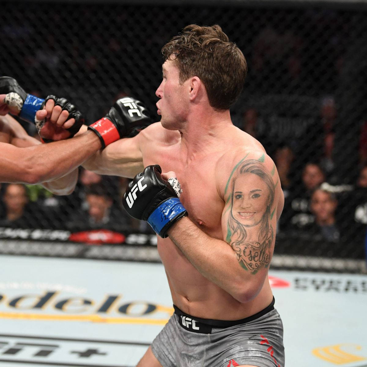 標題：達倫·提爾在高速對決中 壁紙 — 一位肌肉發達的UFC拳手，手臂上有肖像紋身，在明亮的競技場燈光下於八角籠內揮拳。