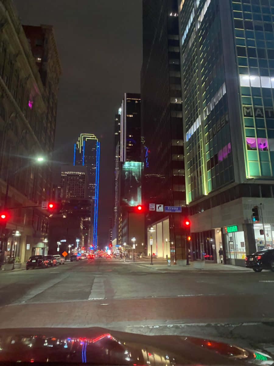 標題：「達拉斯縣日落風景」 壁紙 — 從車內觀看的夜間城市街道景觀，發光的摩天大樓和紅色交通燈照亮潮濕的人行道。