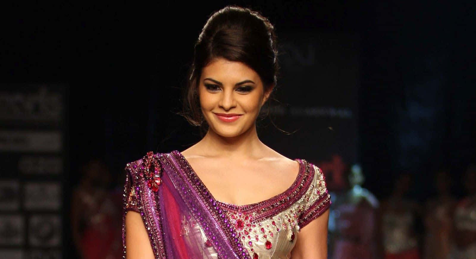 Jacqueline Fernandez 桌布 壁紙 — 模特兒在戲劇性的舞台燈光下，自信地穿著閃閃發光的紫色和銀色刺繡紗麗走秀。