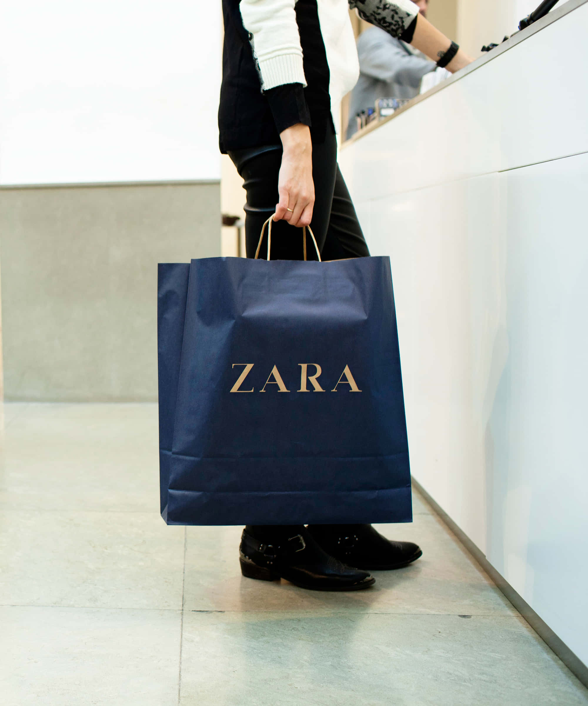 模特兒展現 Zara 最新系列 壁紙 — 一名穿著黑色褲子和靴子的人，在現代零售櫃檯前拿著一個深藍色Zara購物袋。