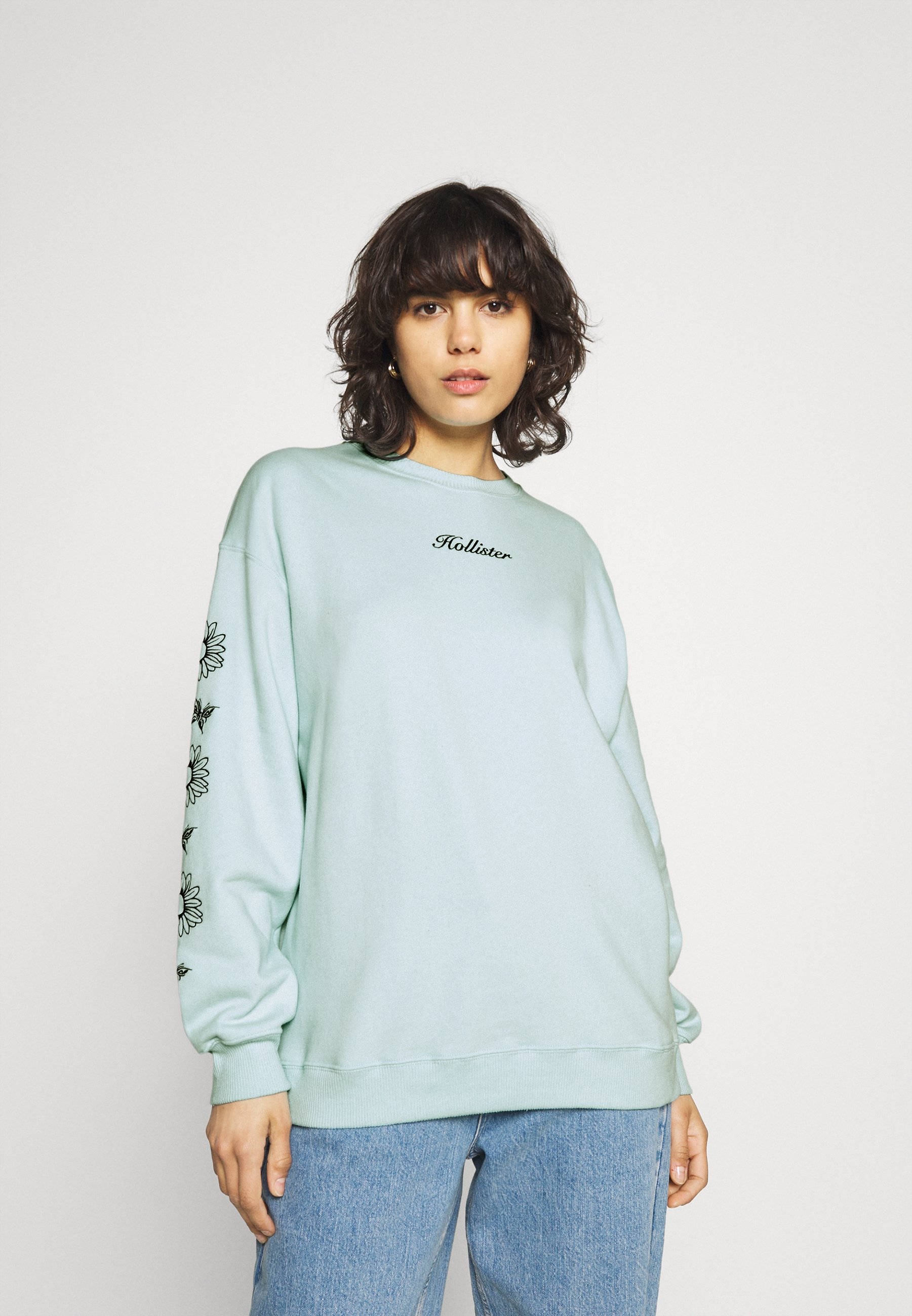 模特身穿 Hollister 衛衣 壁紙 — 一位身穿淺藍色Hollister運動衫、袖子上有花卉刺繡的女性，站在純白色背景前。