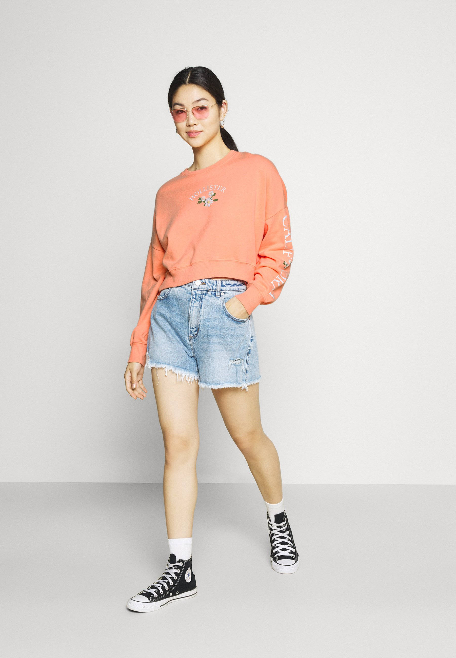 模特身穿桃色 Hollister 衛衣 壁紙 — 一位身穿珊瑚色運動衫和牛仔短褲的年輕女性，在純白色工作室背景前自信地擺姿勢。