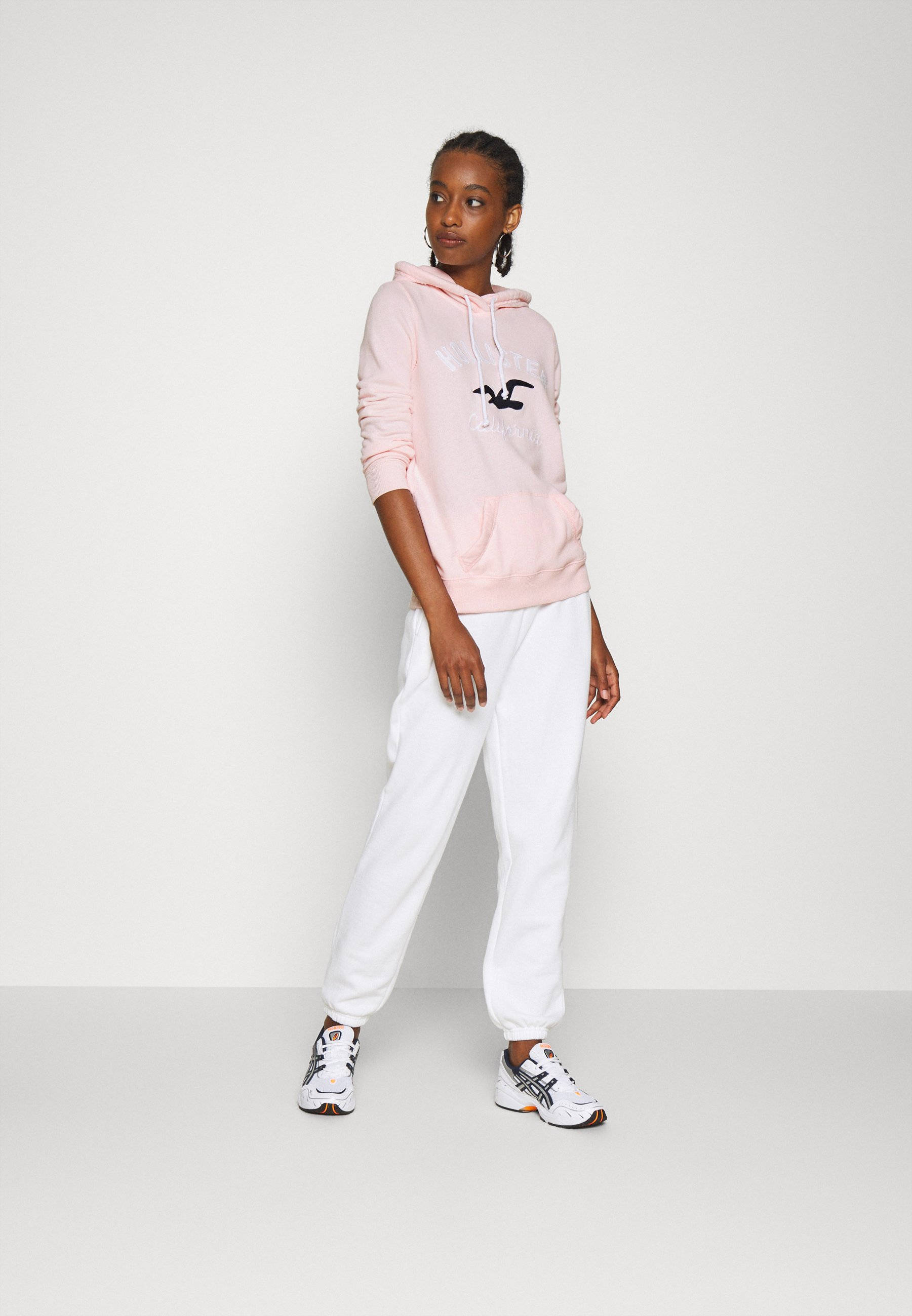 模特身穿粉紅色 Hollister 連帽外套 壁紙 — 一位年輕女性穿著粉紅色連帽衫和白色運動褲，自信地站在純白色工作室背景前。