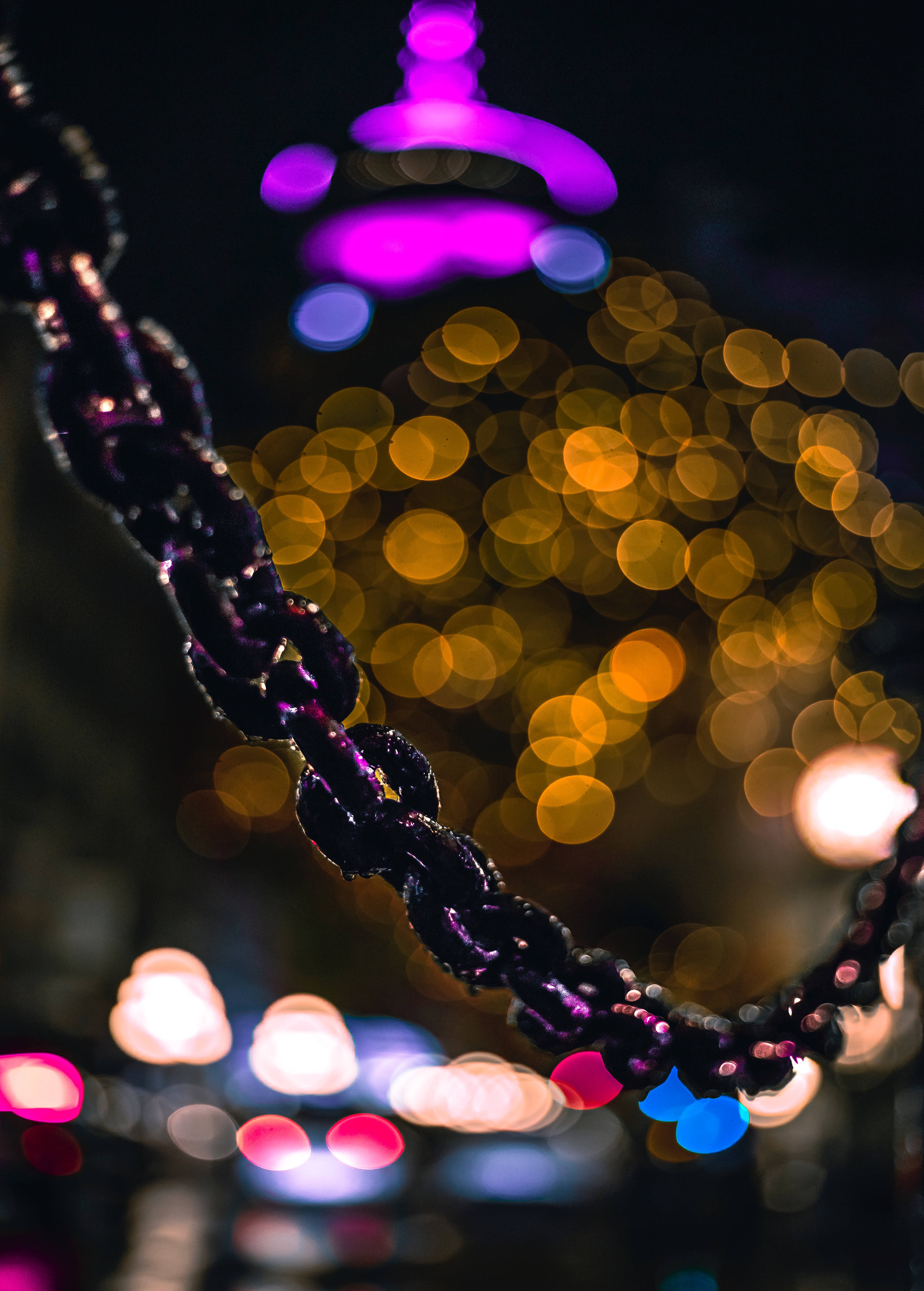 模糊光斑鏈條背景 壁紙 — 夜間暖金色與冷紫色城市燈光散景背景下，帶有紫色高光的深色鏈條特寫。