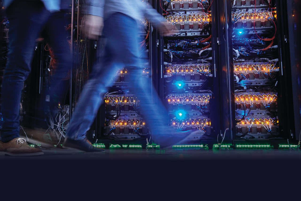 Gaming Servers 桌布 壁紙 — 模糊的技術人員走過昏暗數據中心中發光的伺服器機架，帶有橙色和藍色燈光。