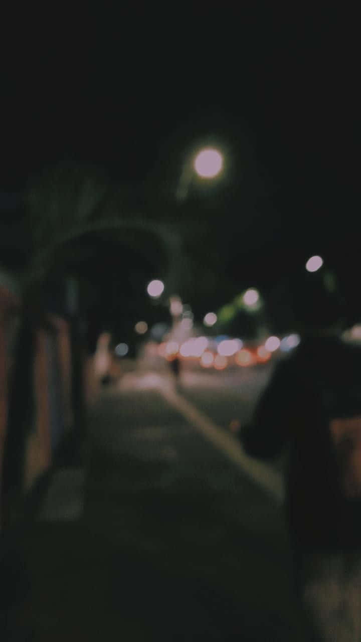 模糊的街燈與汽車畫面 壁紙 — 一個模糊的夜景，一個人走在昏暗的城市人行道上，有散景路燈和遠處的交通。