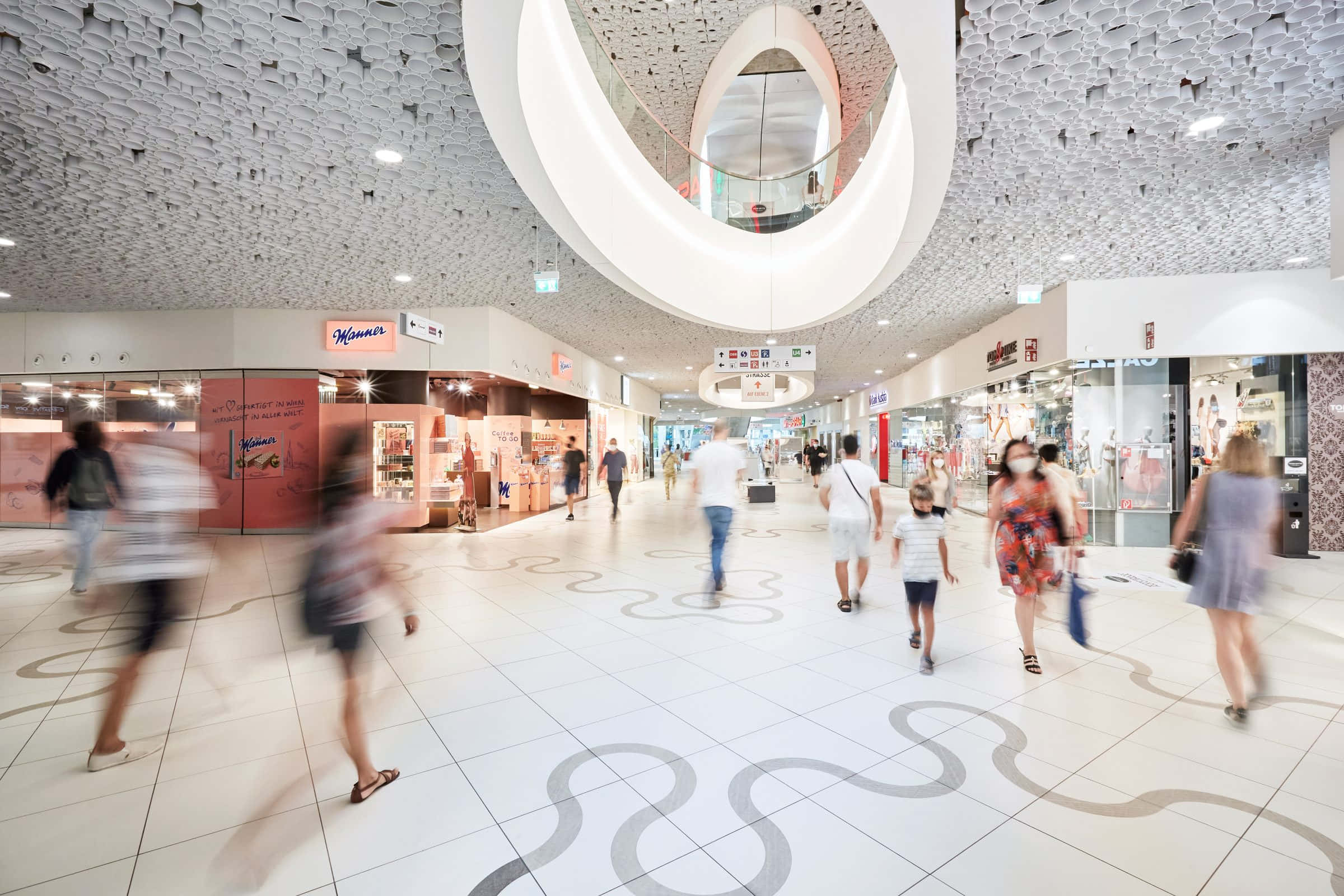 Mall 桌布 壁紙 — 模糊的購物者穿過一個明亮的現代購物中心，擁有雕塑般的白色天花板和圖案地板磚。
