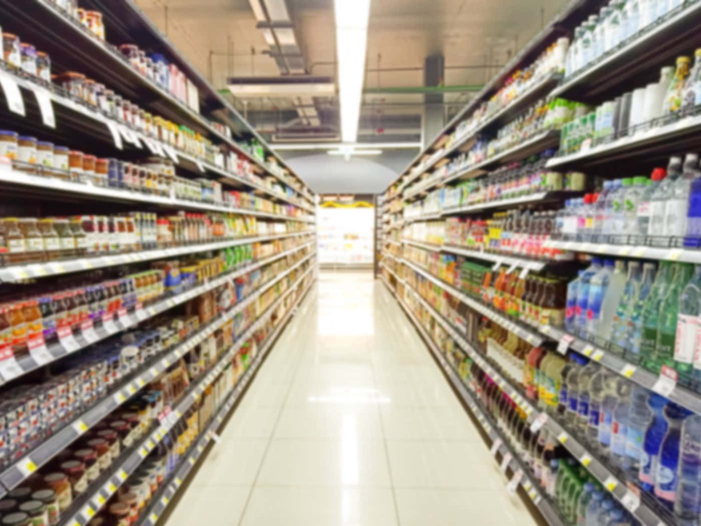 Grocery Store 桌布 壁紙 — 模糊的超級市場通道，貨架上擺滿飲料和罐子，在明亮的螢光燈下。