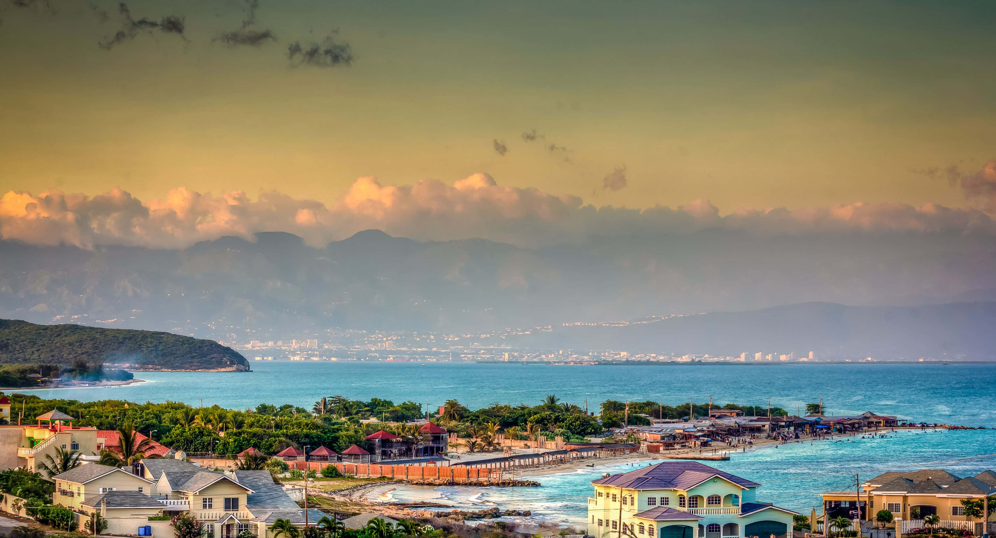 樹木岩石與清澈湖水的寧靜景觀 壁紙 — 沿海小鎮，粉彩房屋和棕櫚樹依偎在碧綠的海水旁，遠方山巒在日落時分。