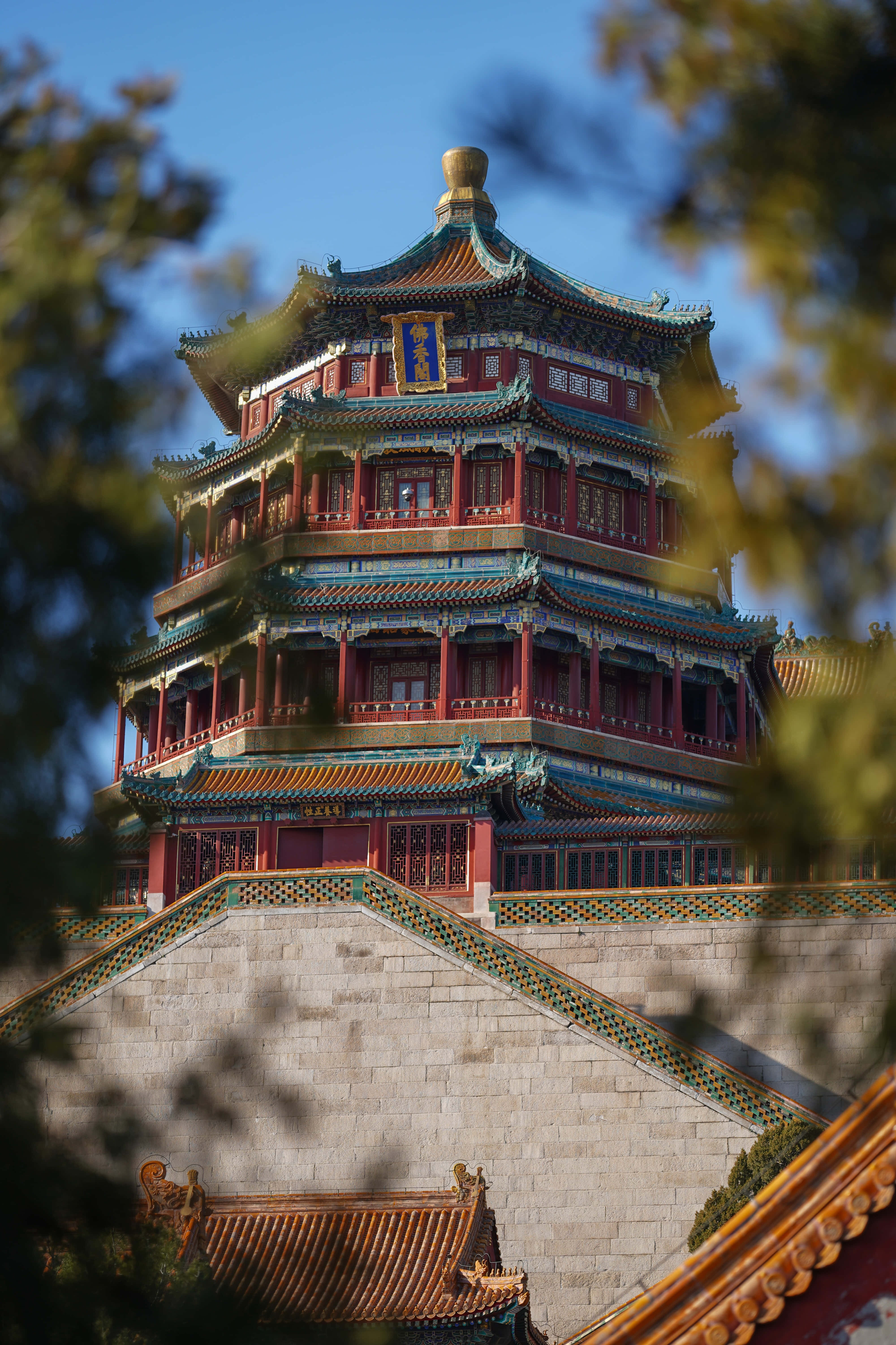 樹林間眺望頤和園 壁紙 — 一座宏偉的傳統中式寶塔，紅牆金瓦，在晴朗的藍天下被模糊的綠色樹葉襯托著。