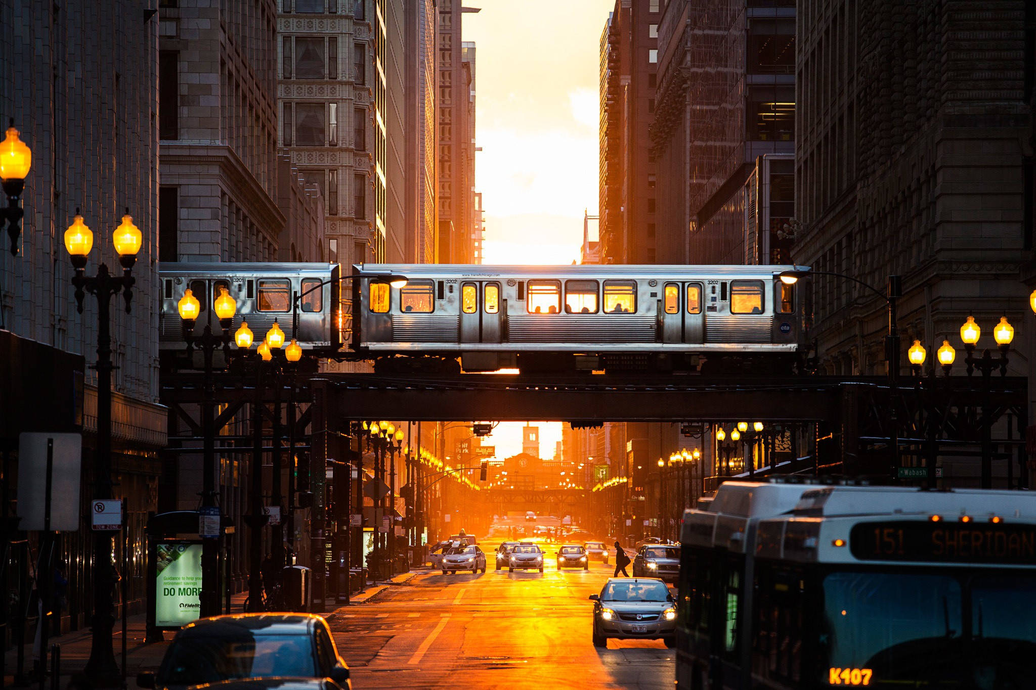 橙城鐵路穿梭繁忙街景 壁紙 — 一列銀色火車在高架軌道上滑行，下方是沐浴在金色夕陽光芒中的繁忙城市街道。