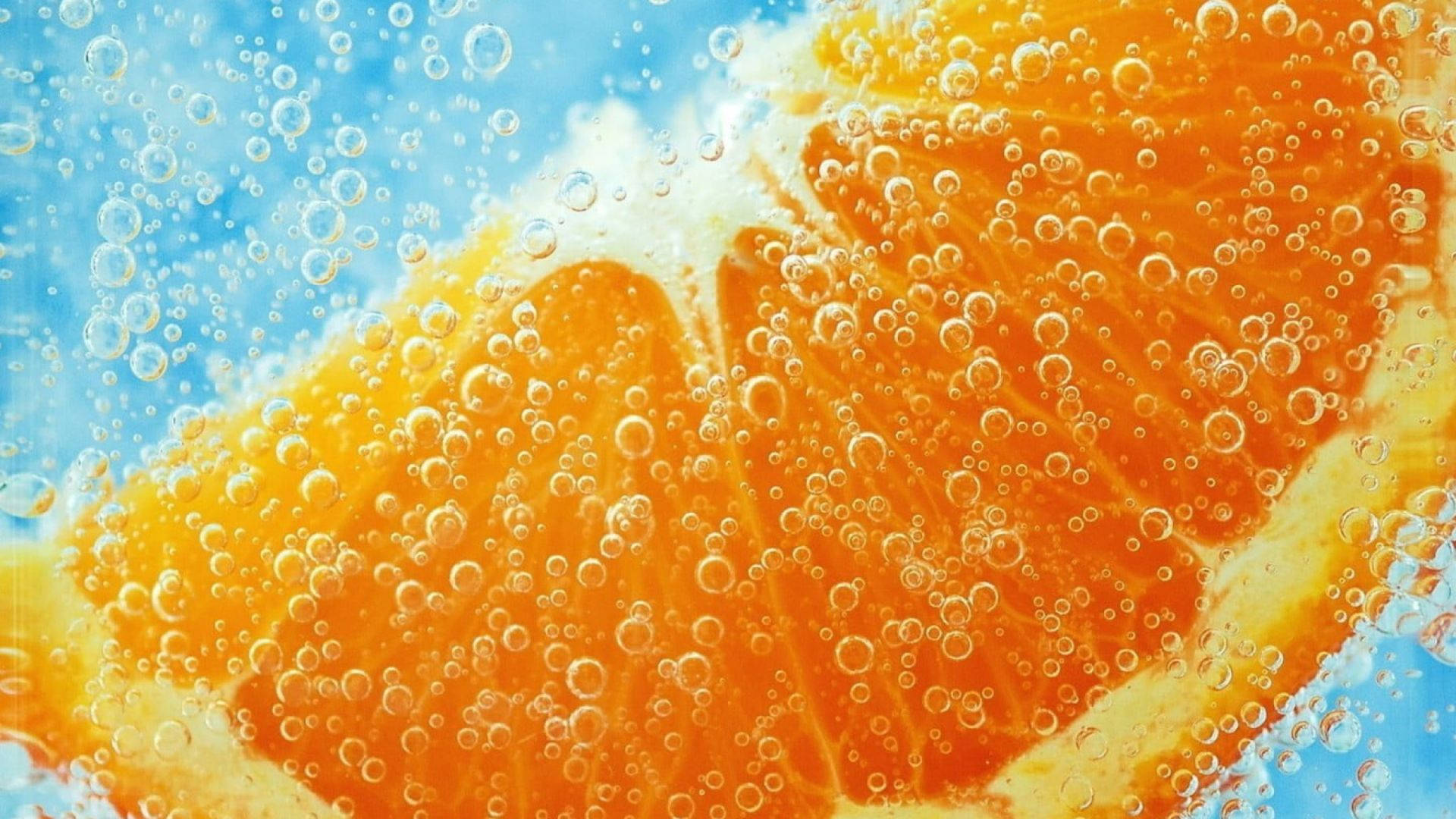 橙子伴氣泡 壁紙 — 鮮豔的橙片浸在氣泡水中，氣泡在明亮的藍色背景上升起。