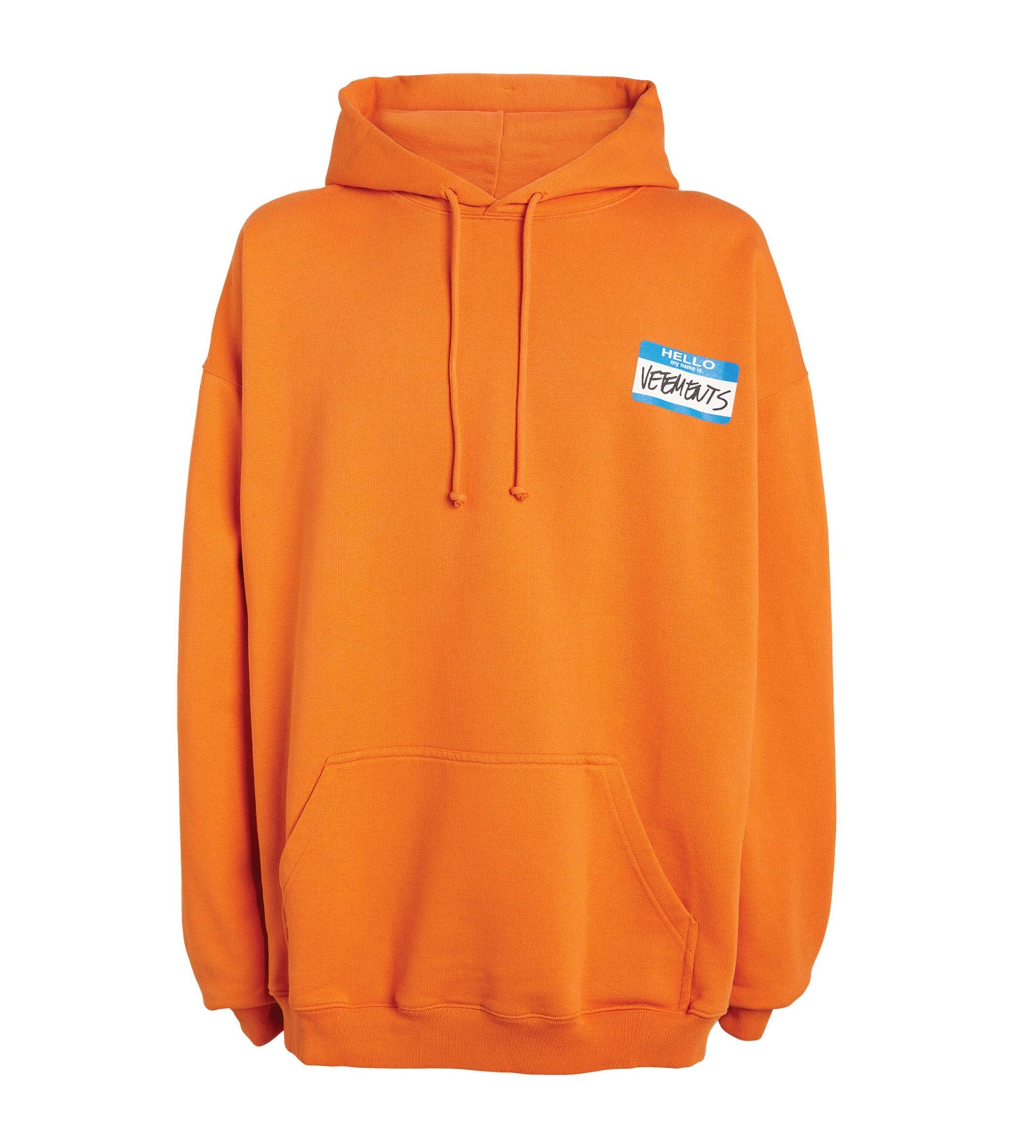 橙色 Vetements 連帽衫 壁紙 — 充滿活力的橙色連帽衫，左胸有一個藍白相間的名牌，上面寫著「HELLO VEEP BUTS」。