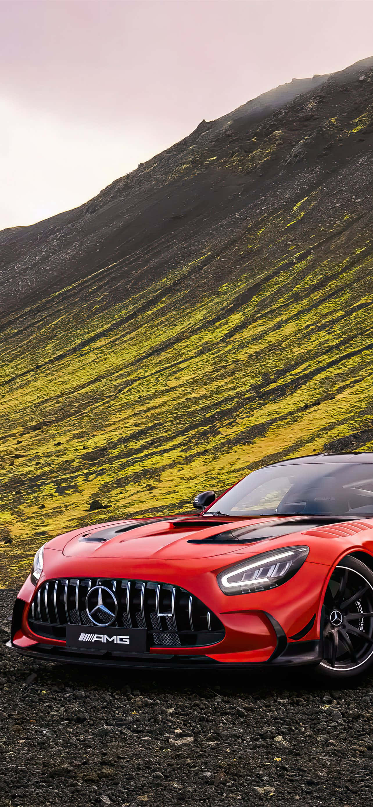 橙色AMG GT-R山丘背景壁紙 壁紙 — 一輛鮮紅色的梅賽德斯-AMG跑車停在碎石路上，背景是陡峭長滿苔蘚的山坡，光線柔和。